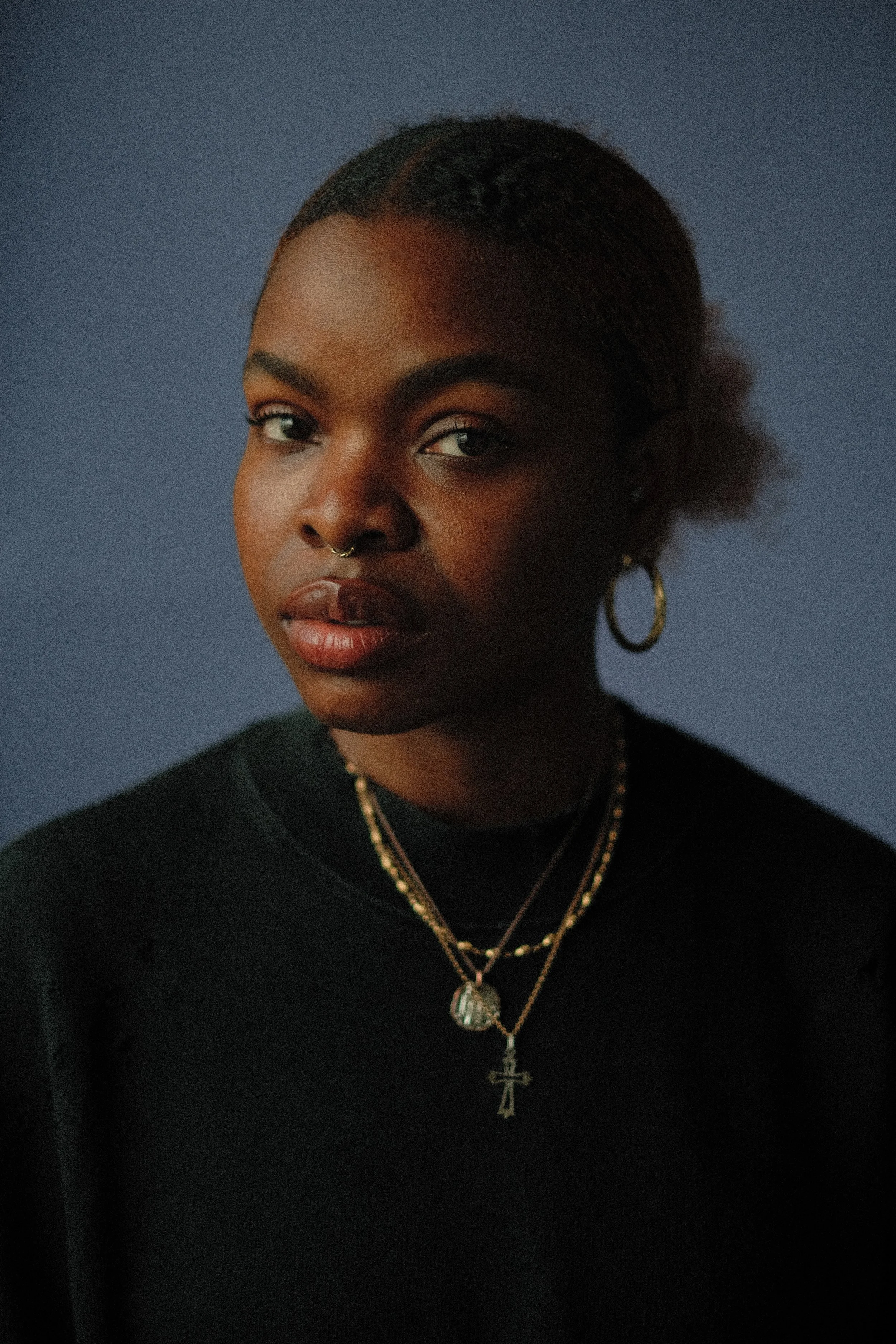 A woman with dark brown hair and light skin sitting and resting her head on her hand, looking to the left, wearing a black blazer over a white top, in a minimalist room.