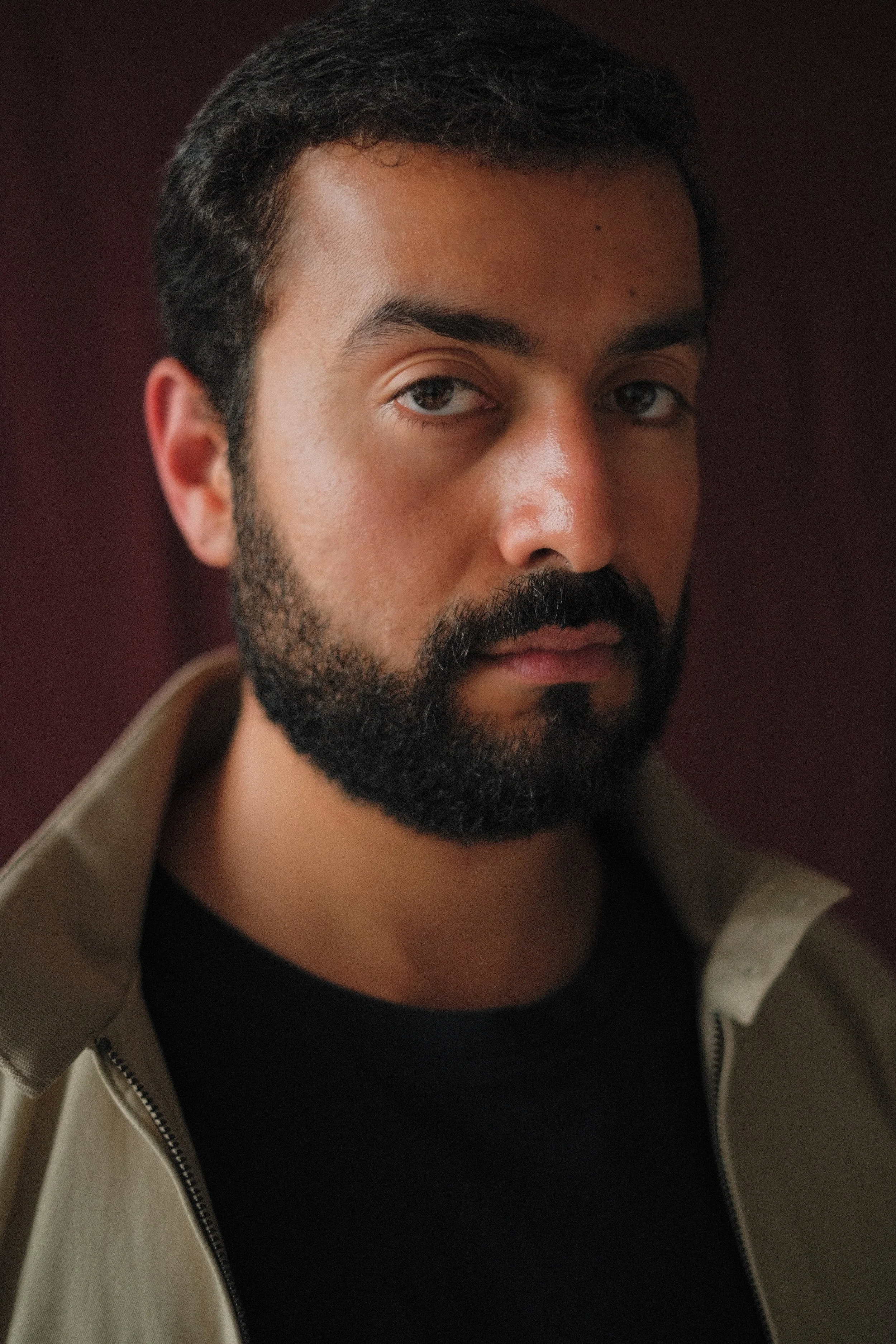 A young man with a short haircut and beard, wearing a black jacket over a white shirt, looking directly at the camera with a serious expression, against a blurred dark pink background.