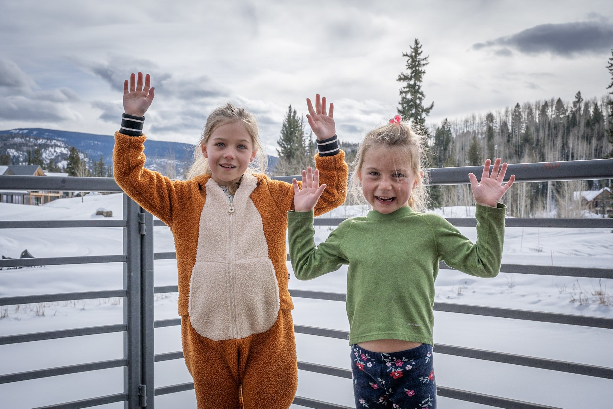 Two young girls standing outside on a snowy day, waving at the camera. One girl wears a brown and beige fleece outfit, the other wears a green long-sleeve shirt and floral pajama pants. They are on a balcony with a snowy landscape in the background and mountains in the distance.