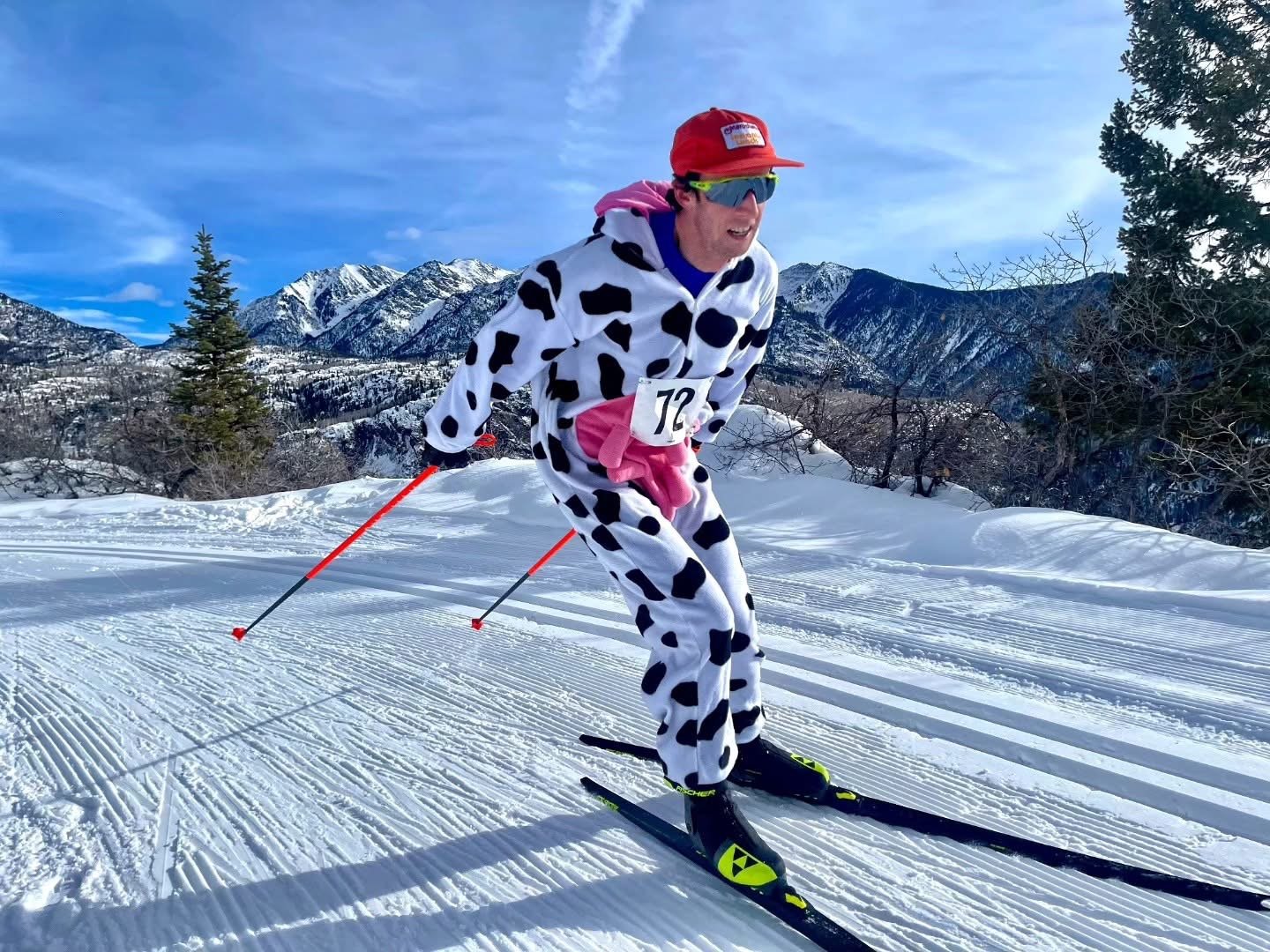 Three people in winter sports attire standing outdoors in snowy mountain landscape. Two adults, a man and a woman, and a young girl. The man is wearing sunglasses, a beige cap, and a white sports bib with the number 55. The woman is wearing sunglasses, a blue cap with Olaf and Durango Nordic written on it, and a white sports bib with the number 69. The young girl wears sunglasses, a hooded jacket, and a backpack. All are smiling and holding ski poles.