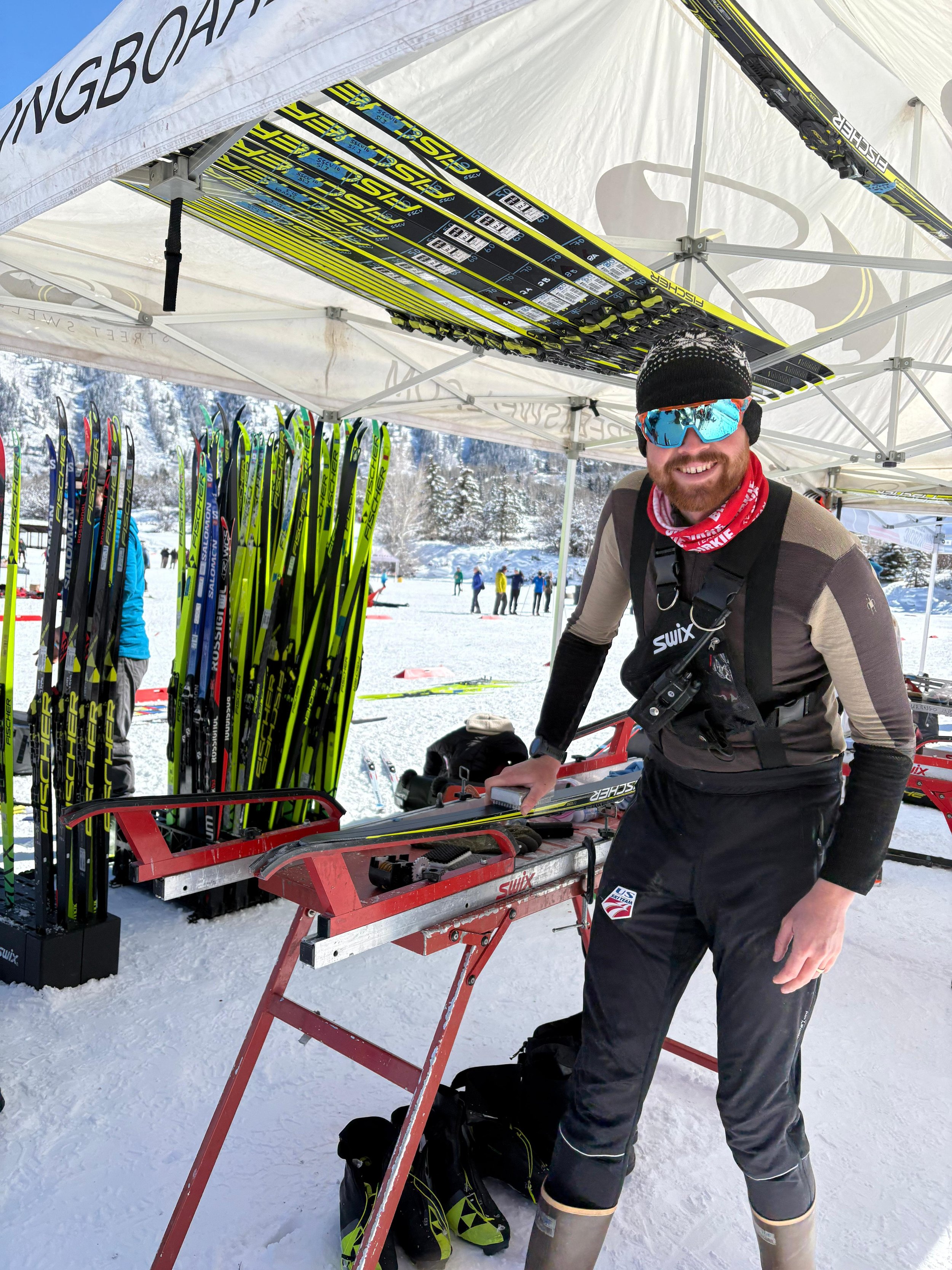 A man with a beard smiling outdoors, wearing a helmet, a bandana, and a backpack, holding a ski pole, with a background of trees and mountains.