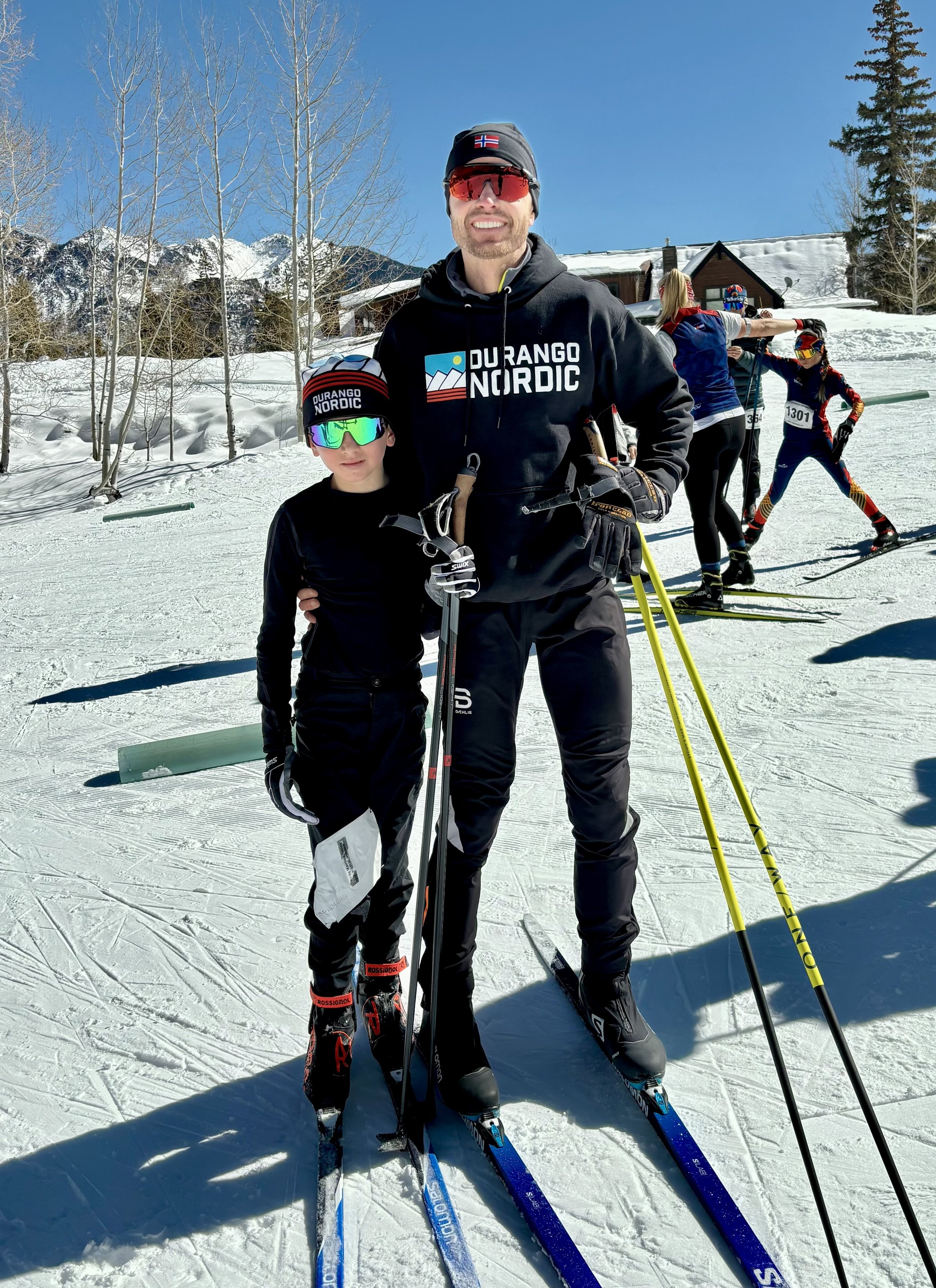 Portrait of a man with a beard and a checkered shirt, smiling, standing against a plain wall.