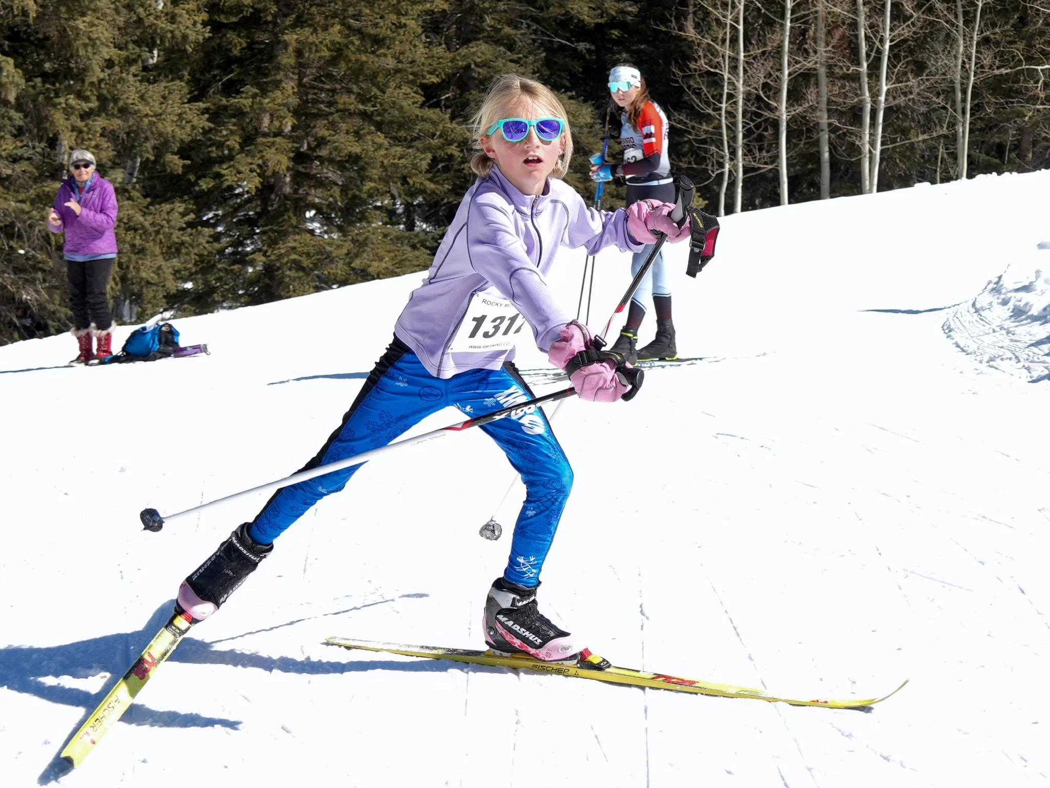 A young girl skiing on snow, wearing blue ski gear, pink gloves, sunglasses, and a race number 131, with two other people in the background on a snowy hillside surrounded by trees.