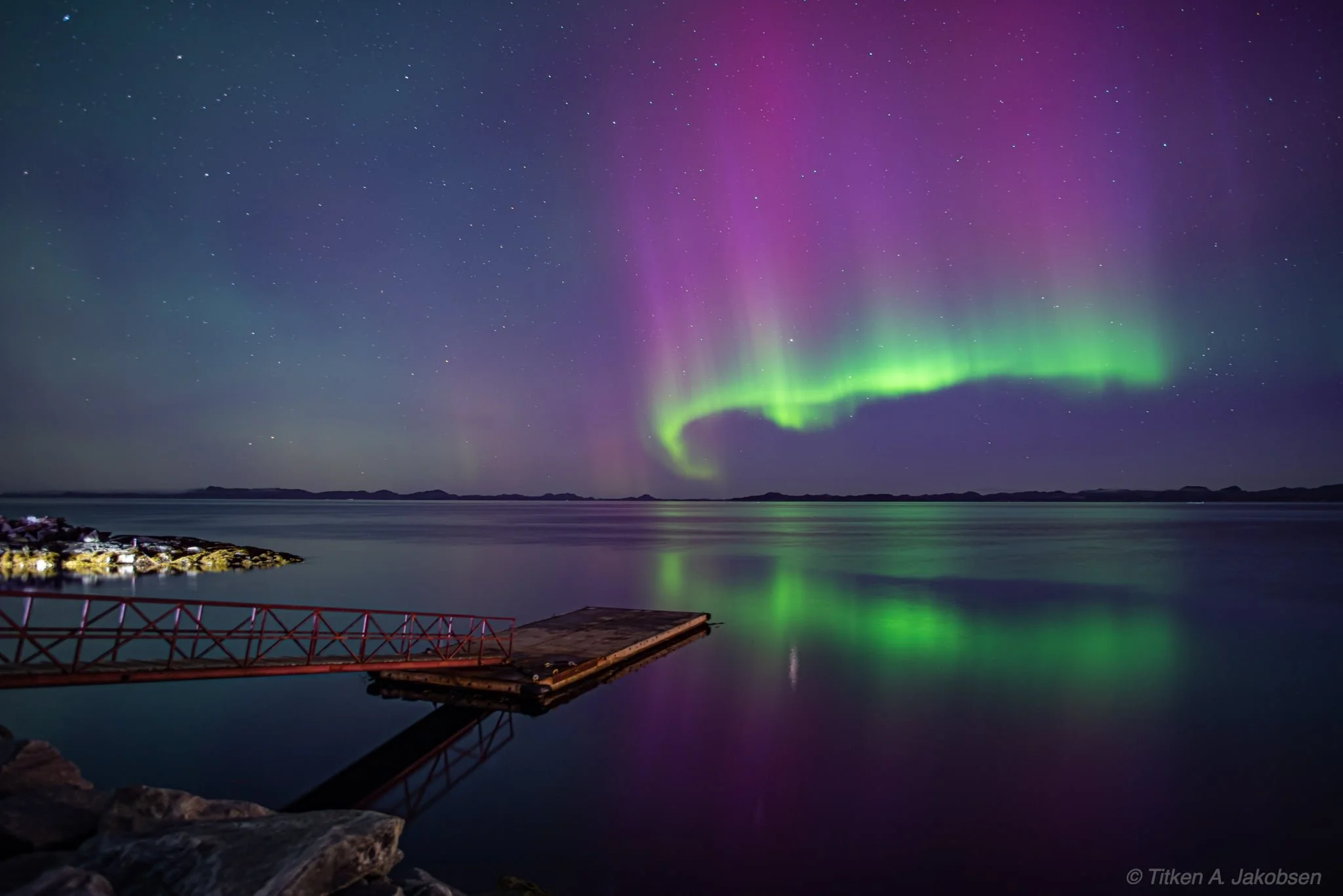 Northern lights over a calm body of water with a small dock, rocky shoreline in the foreground, and distant land on the horizon.