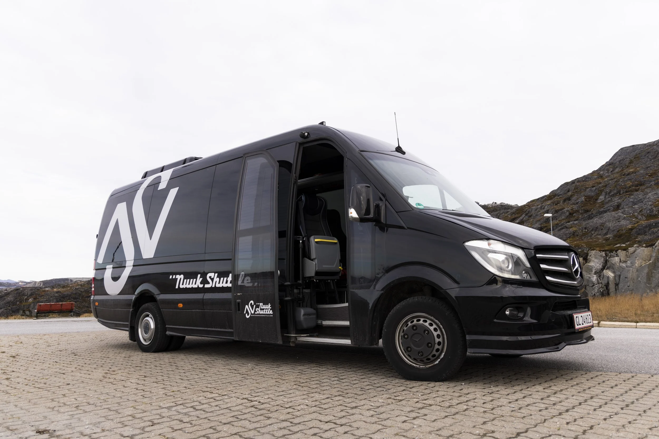 Black Mercedes-Benz van parked on paved area with rocky hills in background, open door showing interior seats