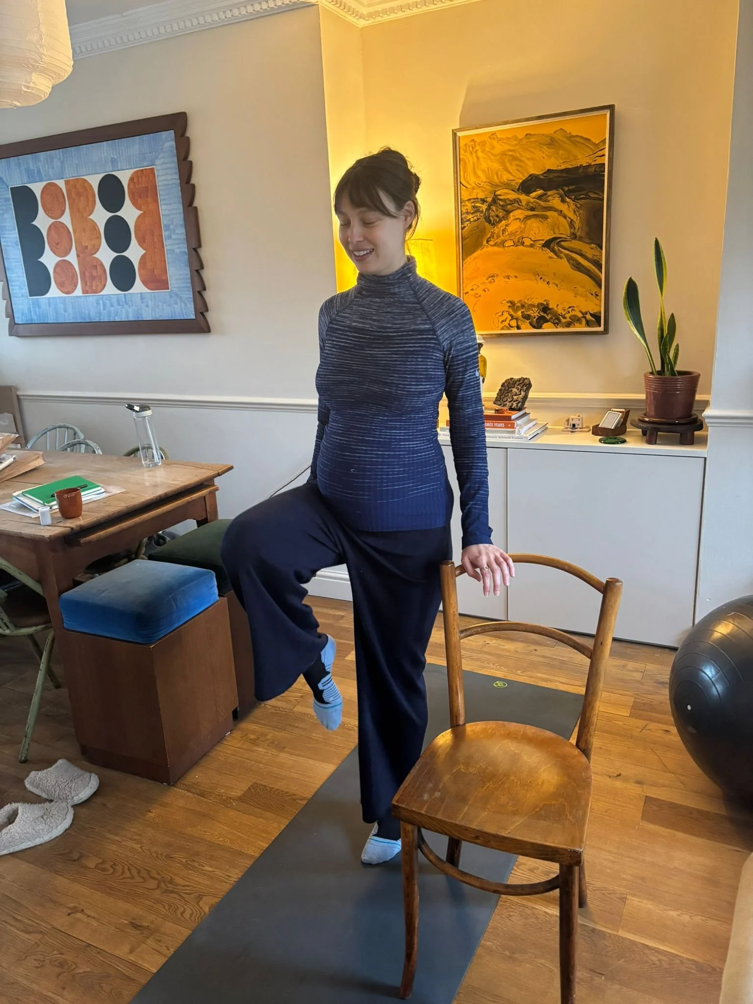 A woman is balancing on one leg with her foot resting on a wooden chair, in a living room.