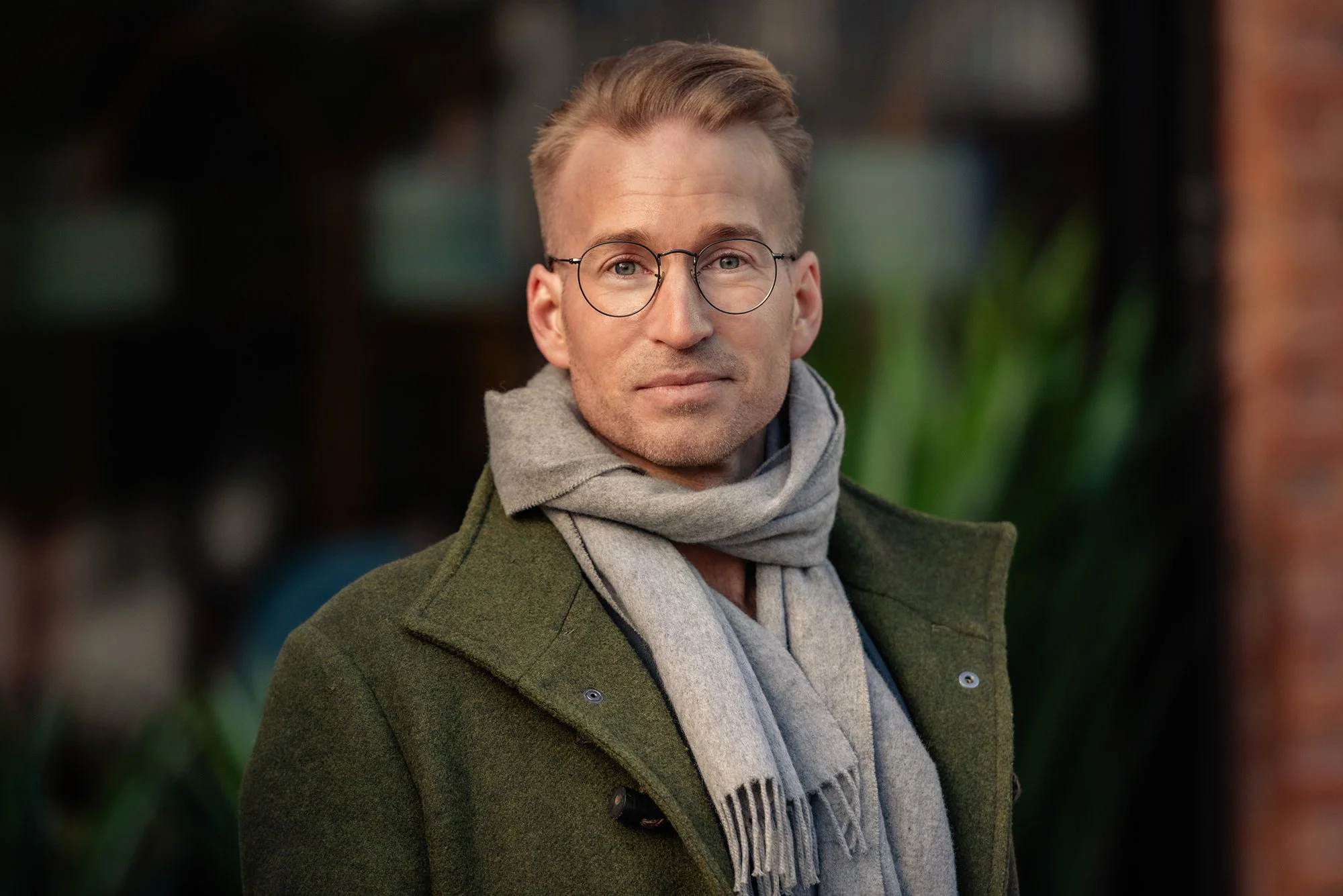 En mann med lys hud, kort blond hår og runde briller, iført en grønn frakk og en grå skjerf, står utenfor med en bakgrunn av grønne planter og en murvegg.