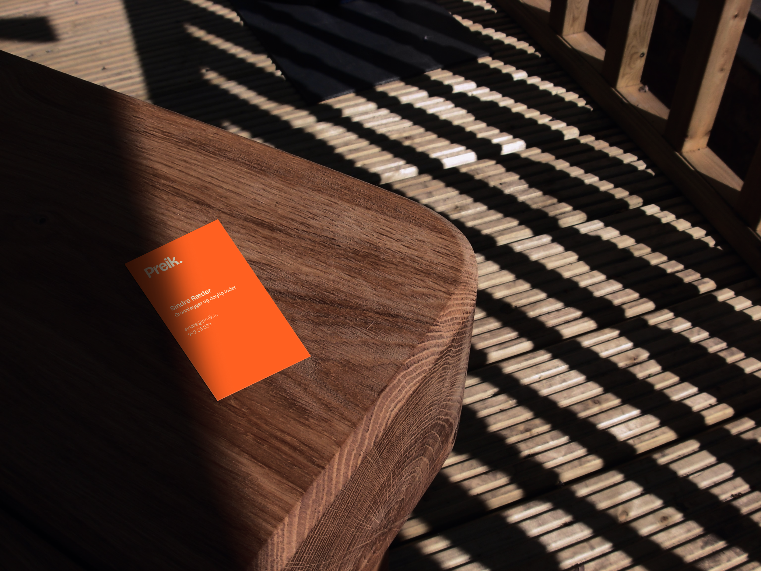En orange visittkort liggende på et trebord. Bakgrunnen består av en terrass med parallelle trelister og skygger.