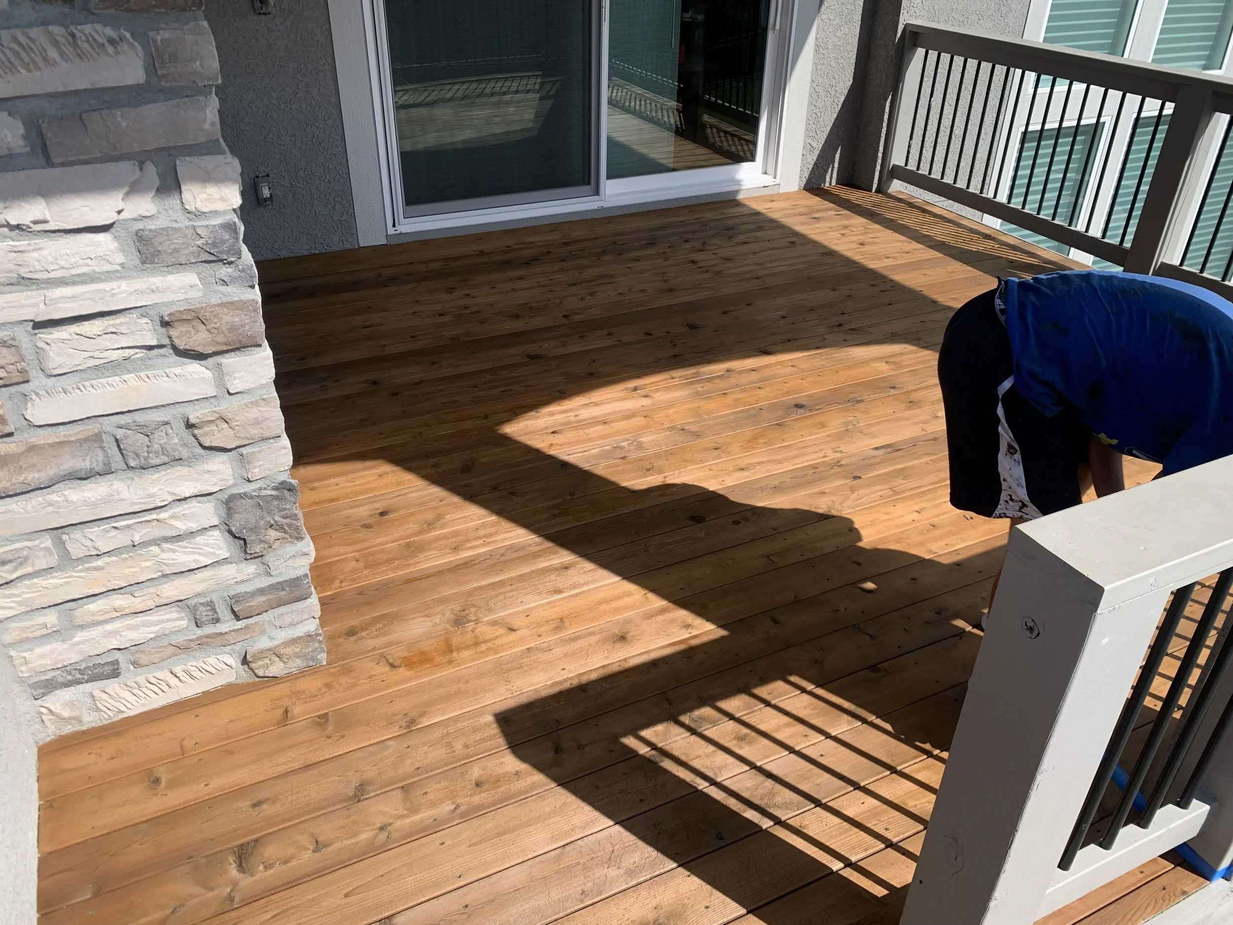 Newly stained wooden porch with a person working near the railing, adjacent to a sliding glass door, with a brick accent wall and apartment complex in the background.