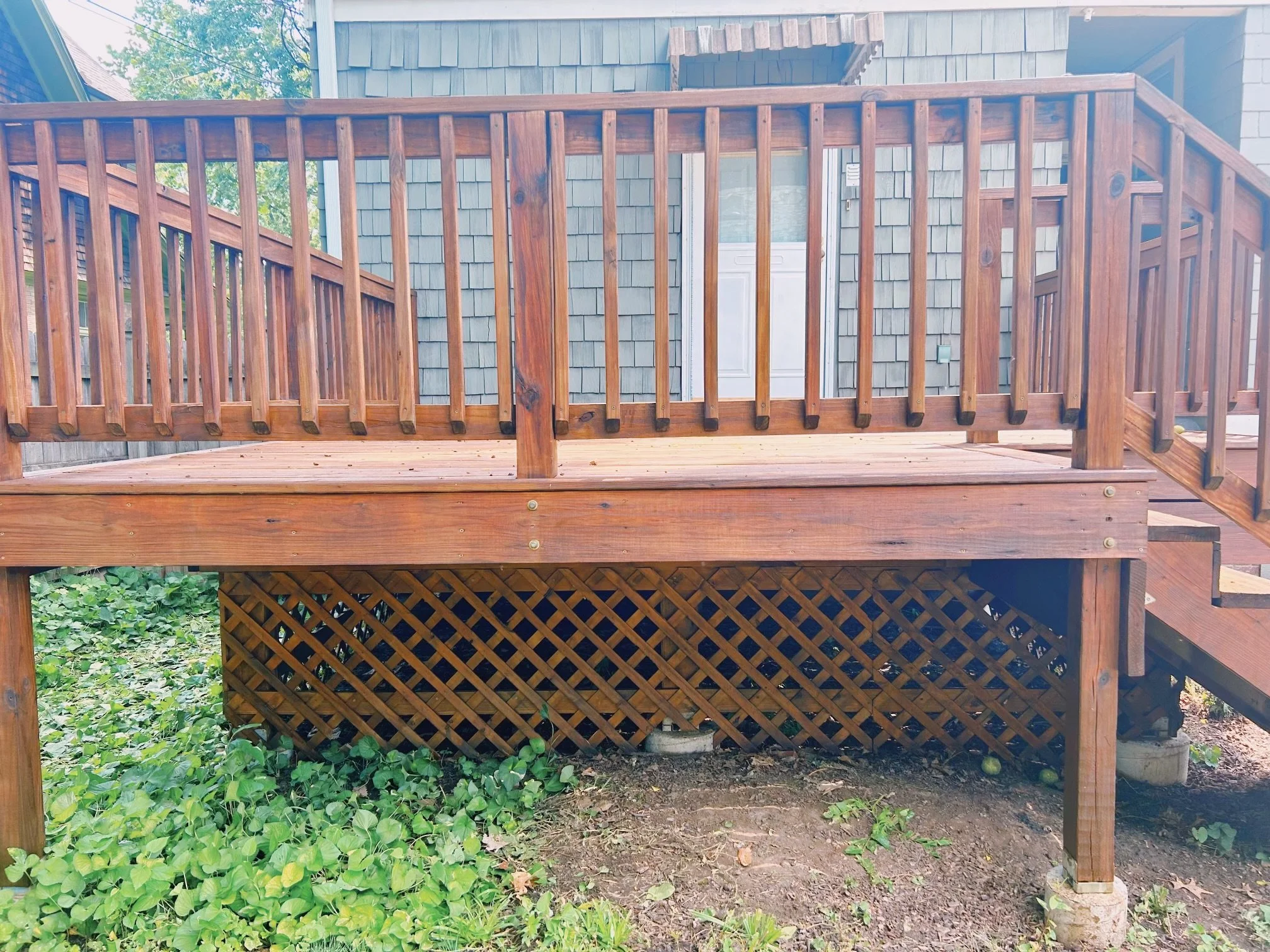 Wooden deck with railing and stairs, attached to a house with blue siding and shingles, with greenery and ground below.