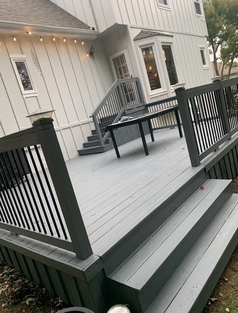 Exterior view of a gray wooden deck with stairs and black railings, attached to a white house with multiple windows and a door, with string lights hanging on the house.