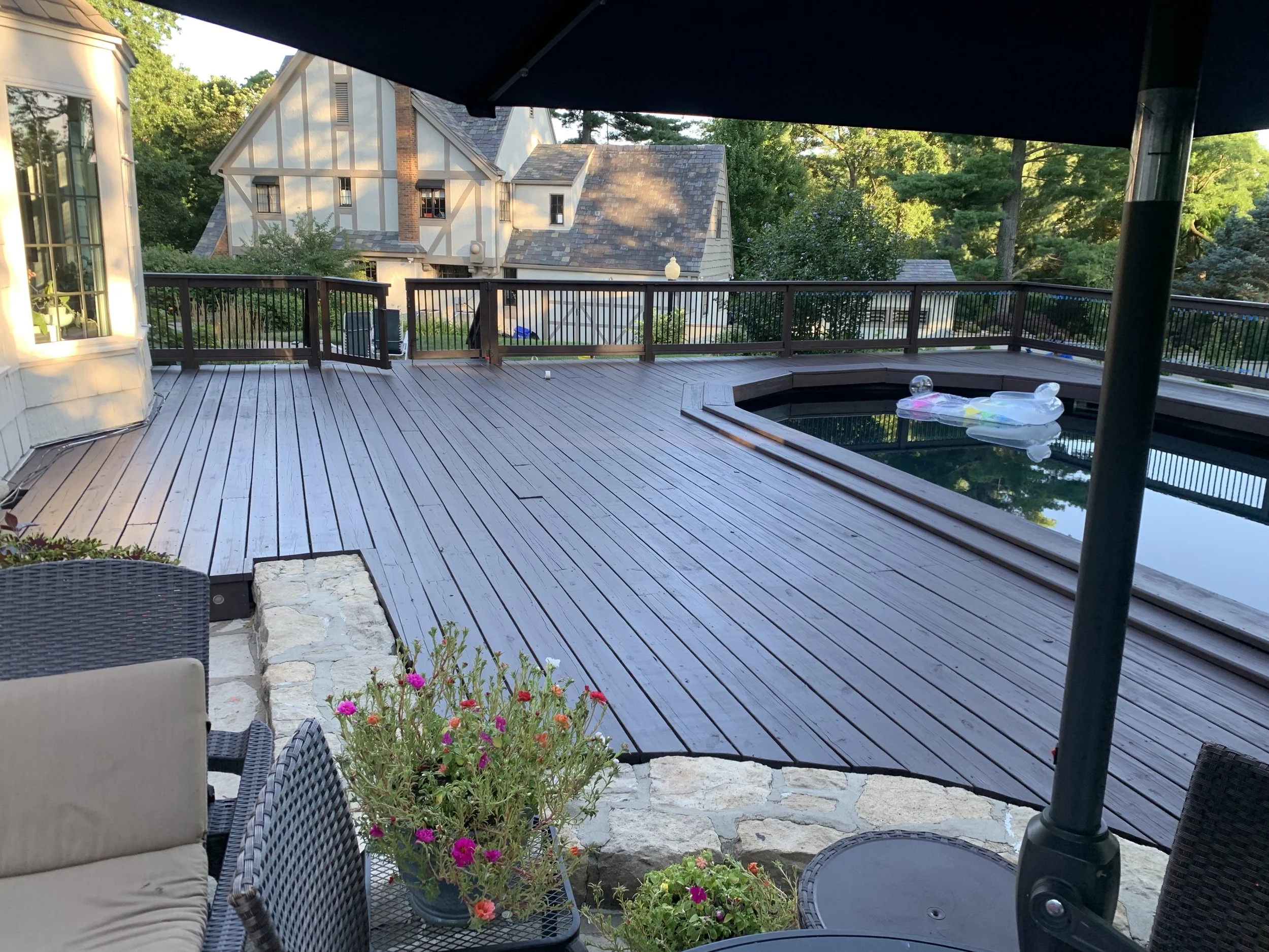 Wooden backyard deck with hot tub, patio furniture, potted flowers, and a large patio umbrella, overlooking neighboring houses and trees.