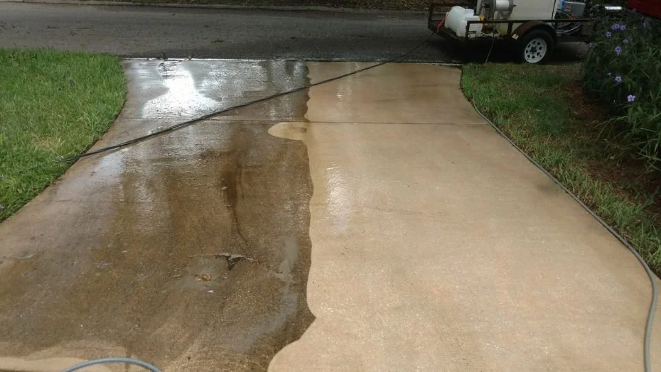 A concrete driveway is being pressure washed, showing a clean, washed side on the right and a dirty, streaked side on the left, with a garden hose and a pressure washer in the background.