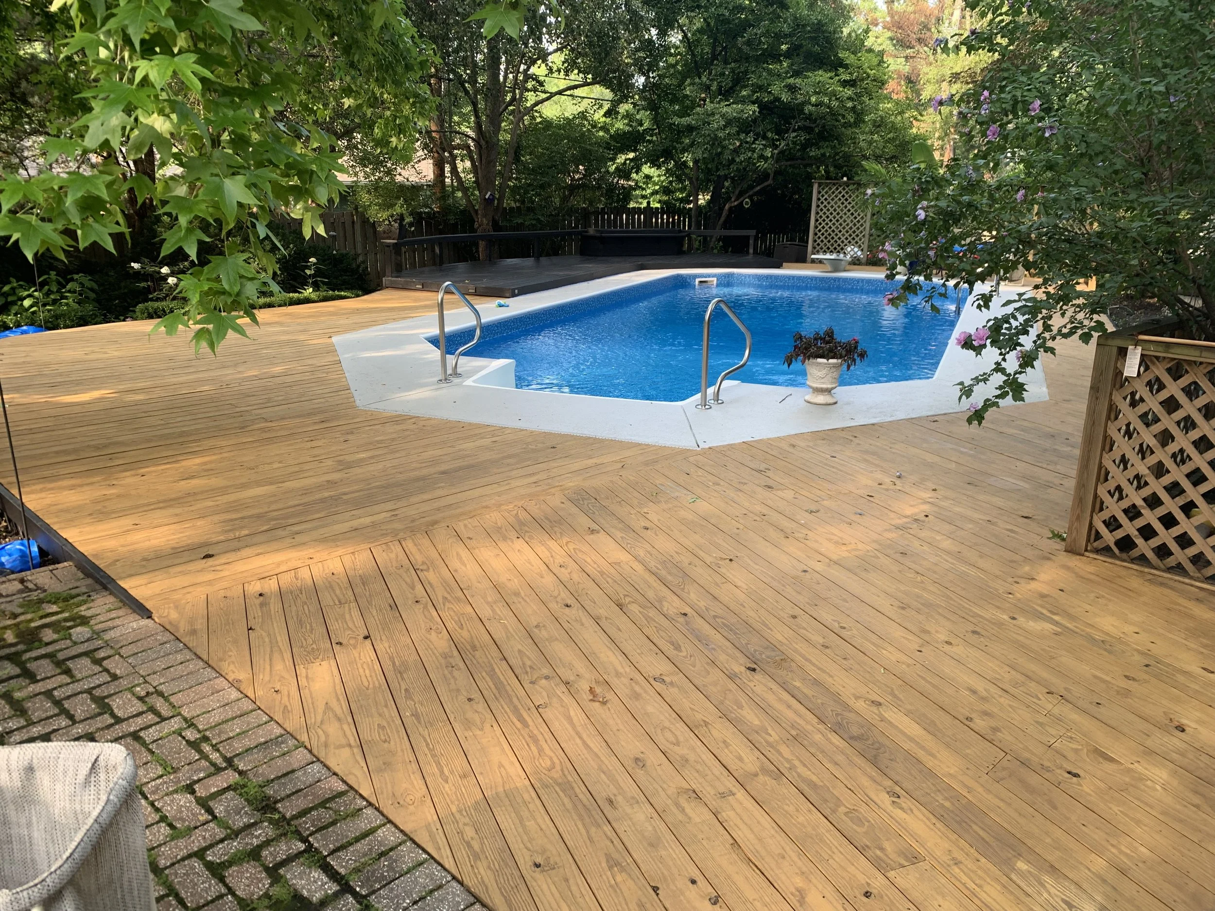 Newly constructed wooden deck surrounding a blue swimming pool in a backyard, with trees and bushes providing privacy.