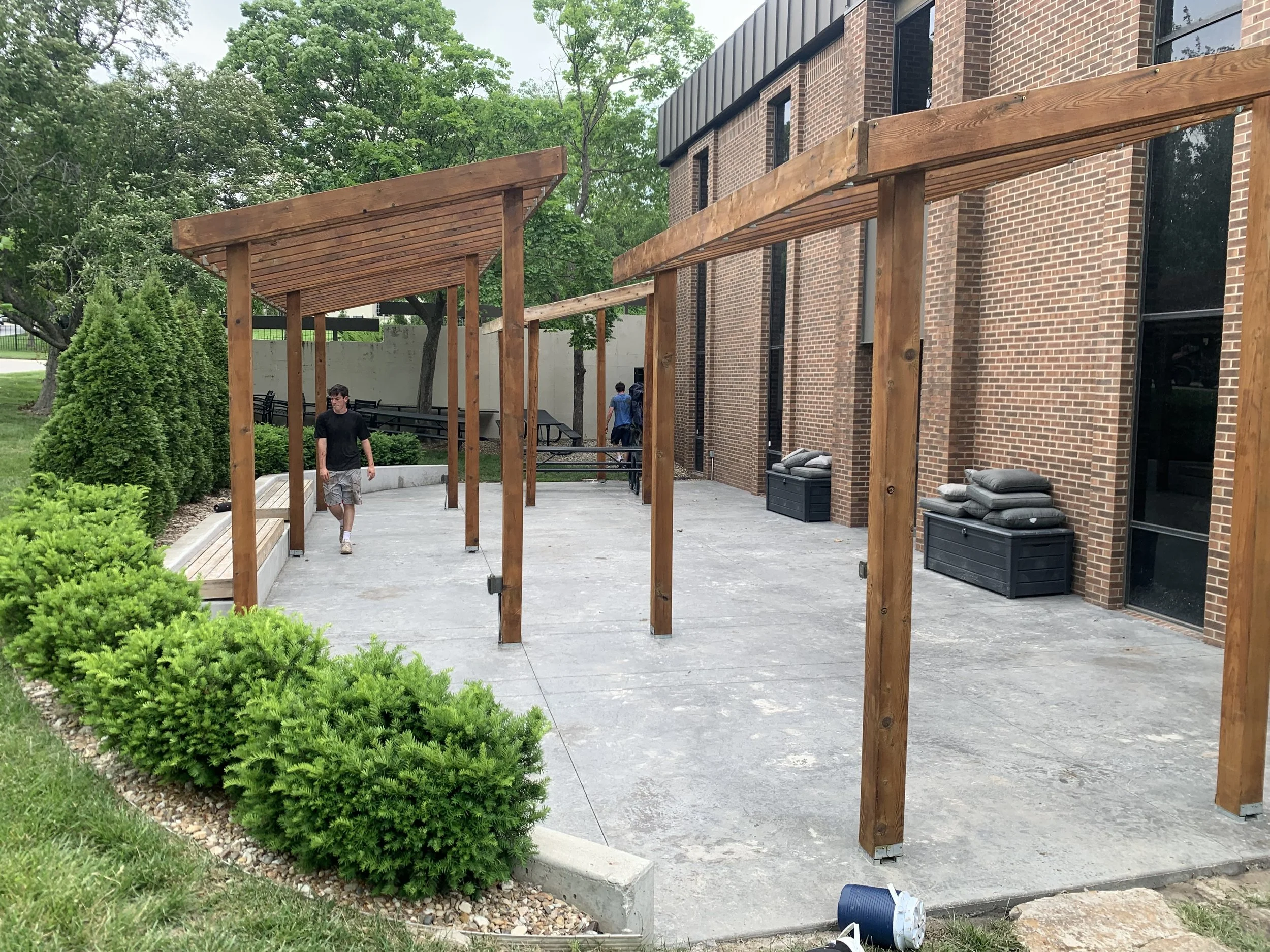 A brick building with a concrete patio under construction, featuring a wooden frame for a future pergola, and a man walking on the patio. There are bushes and trees surrounding the area.