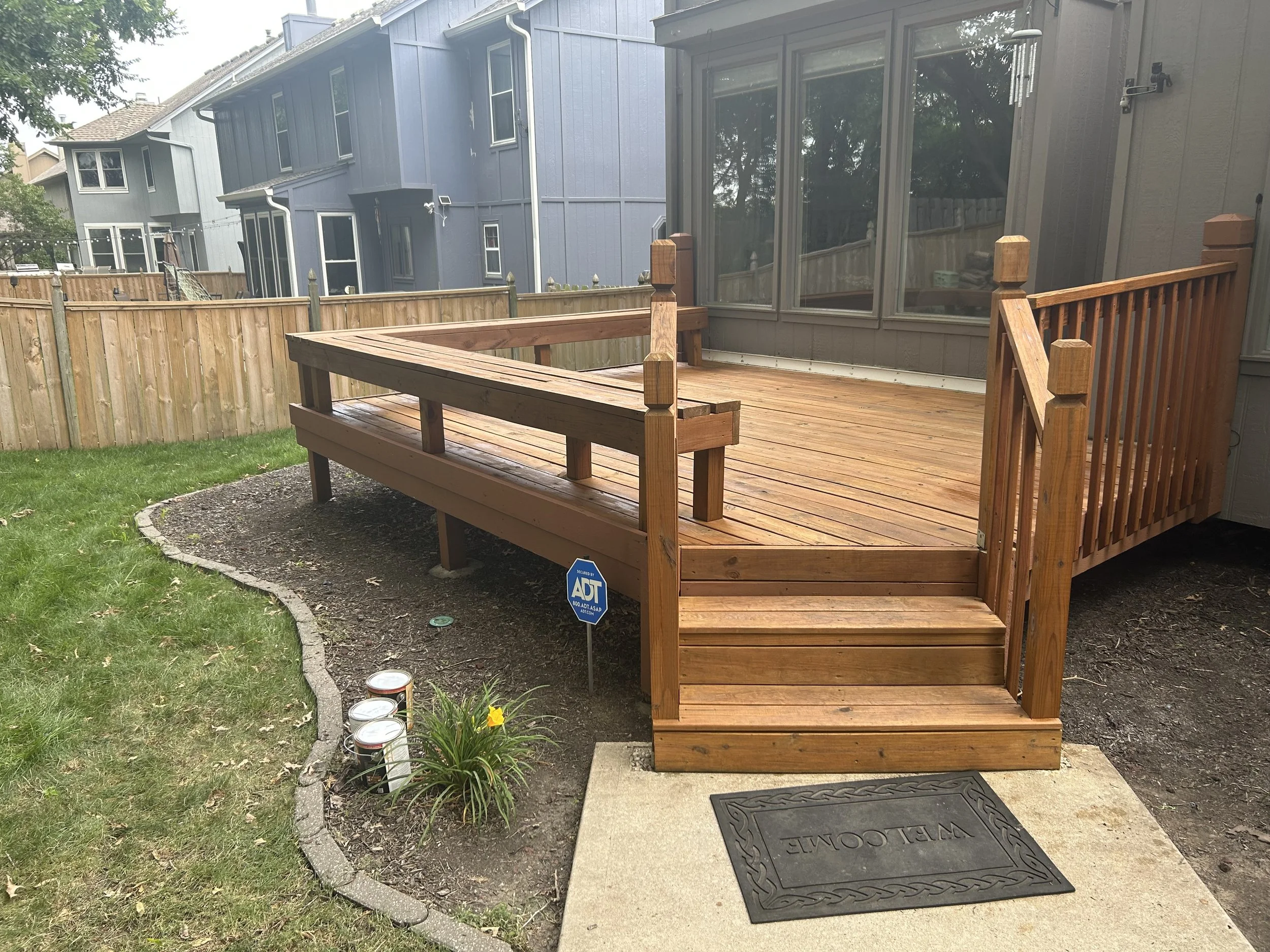 A newly built wooden deck with steps leading up to it, attached to a house, in a backyard with a grass lawn and a wooden fence. There is a Welcome mat at the steps and a small flower bed with plants and an ADT security sign.