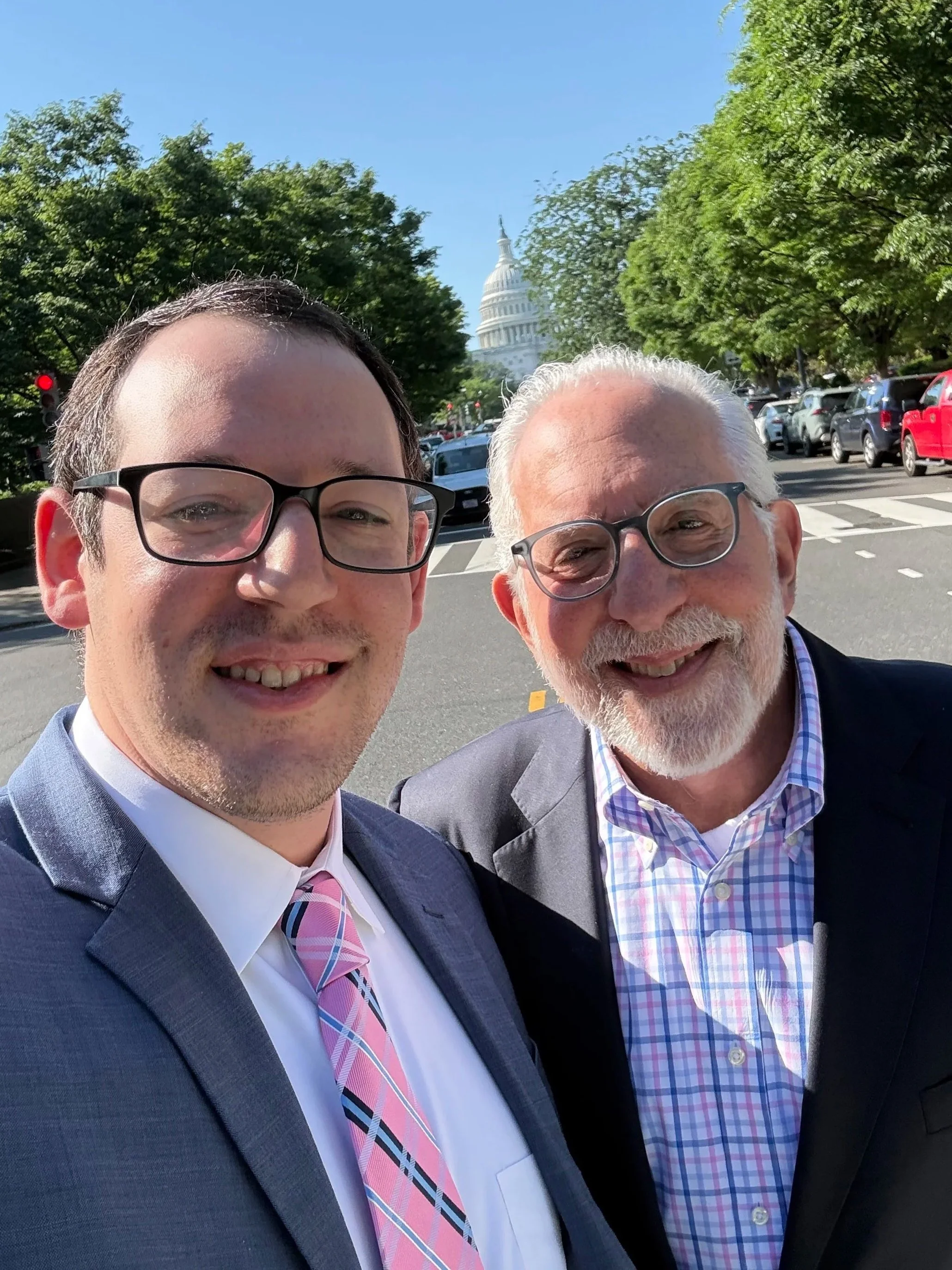 Close-up of two smiling men in suits taking a selfie outdoors with the U.S. Capitol building and rows of parked cars in the background on a sunny day.