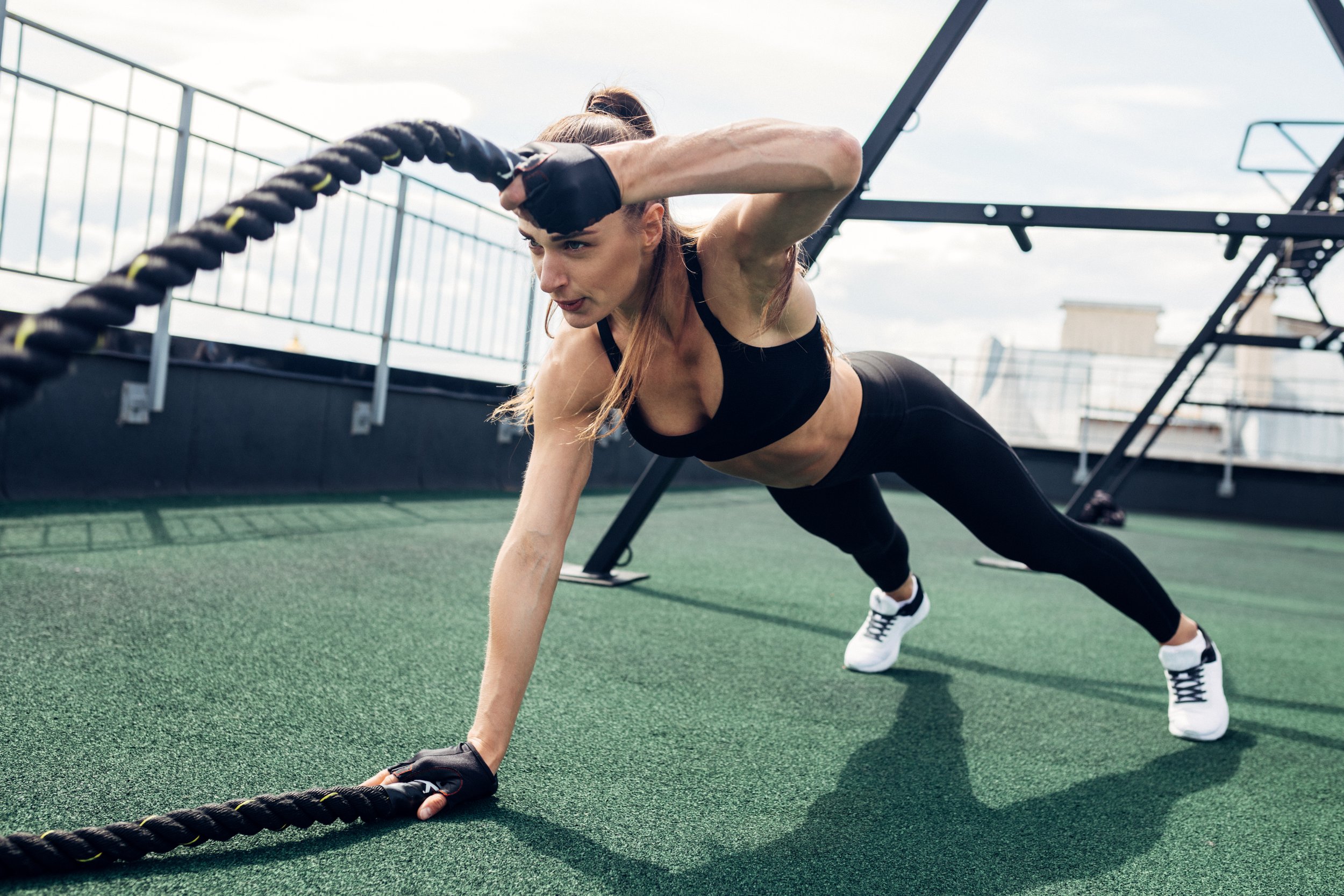 A woman in athletic clothing and gloves exercises on a rooftop gym, doing a plank with one arm on a battle rope, with city buildings in the background.