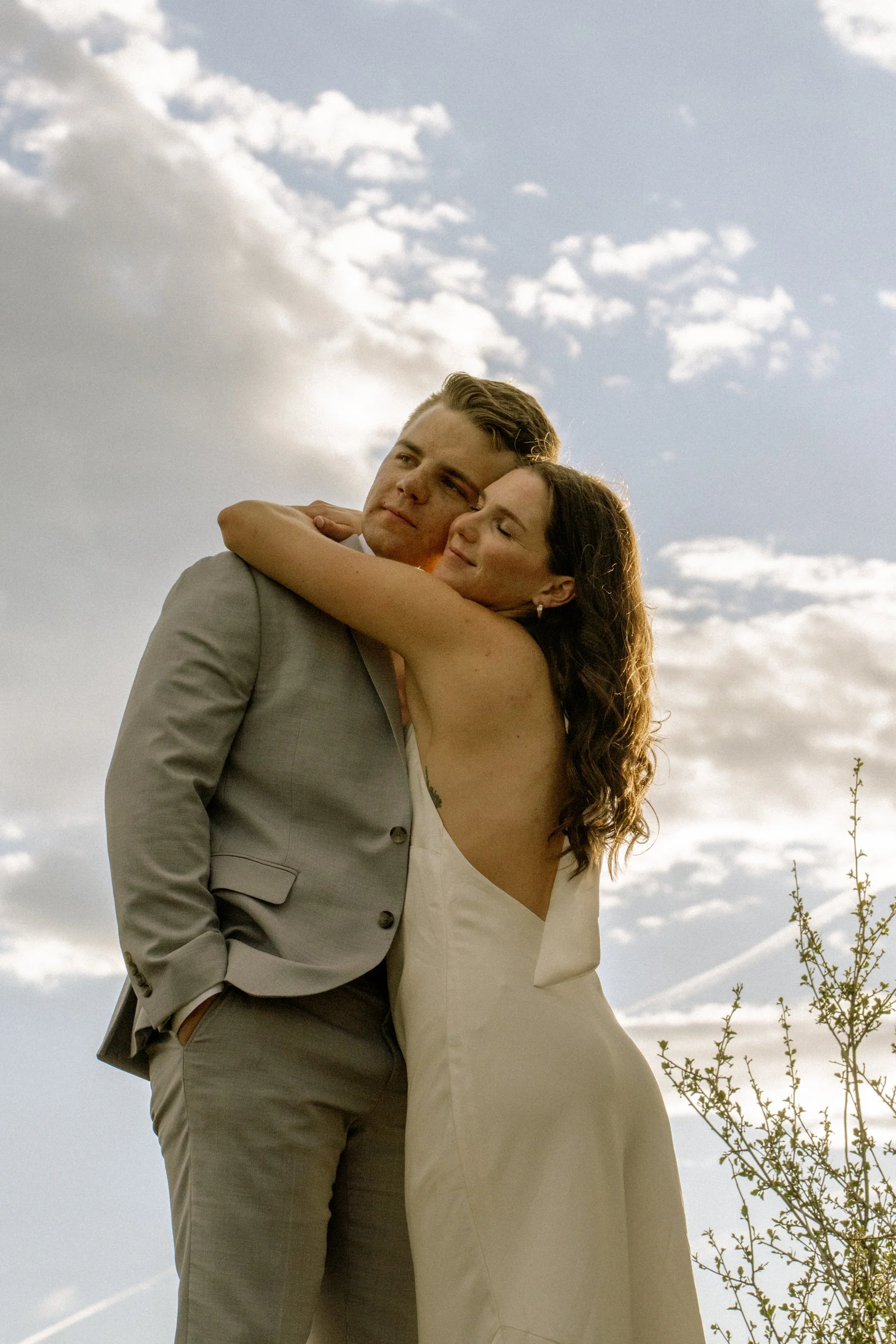 A man in a light gray suit and a woman in a white dress embracing outdoors under a cloudy sky.