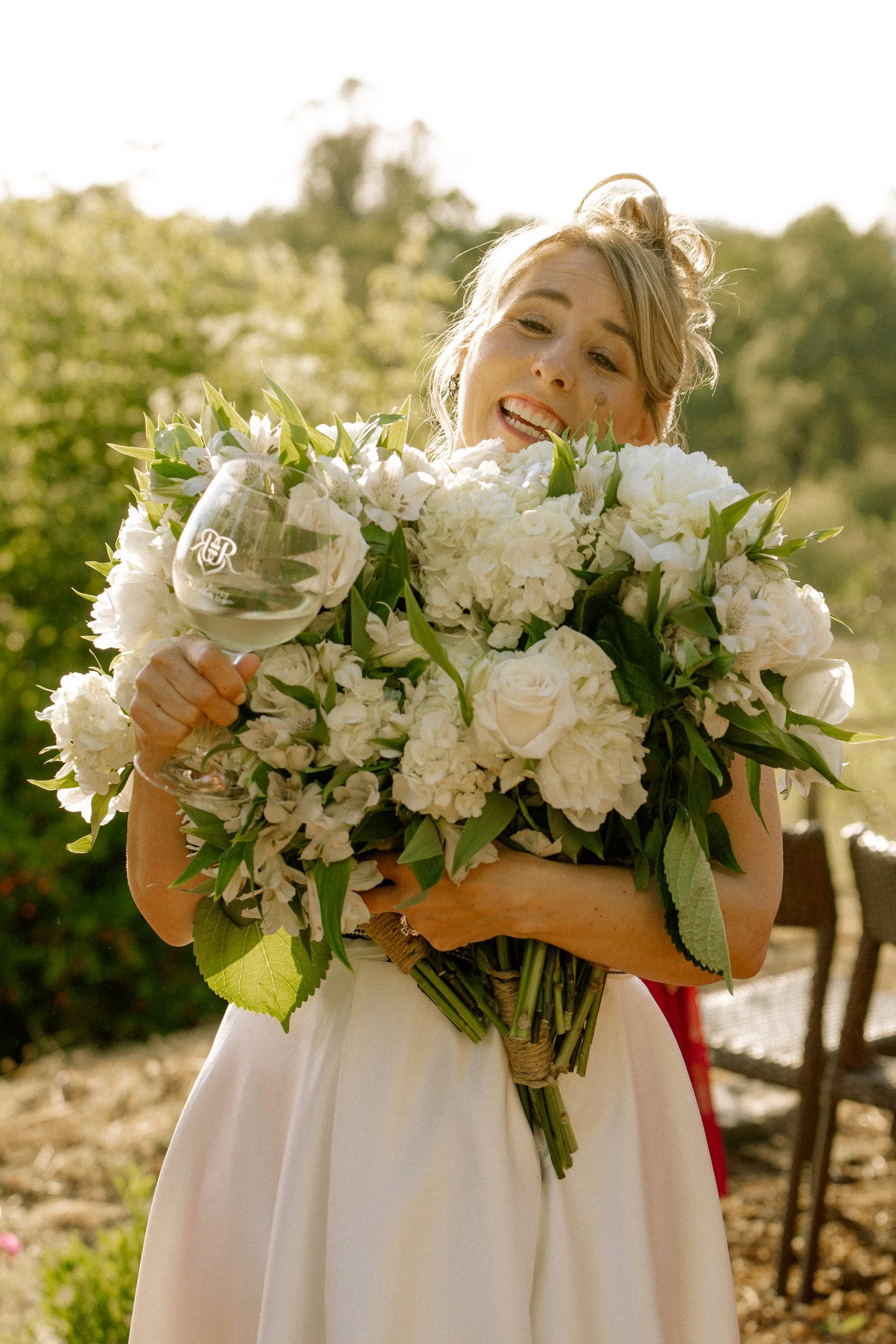 A smiling bride holding a large bouquet of white flowers and a wine glass, outdoors in bright sunlight with blurred trees in the background.