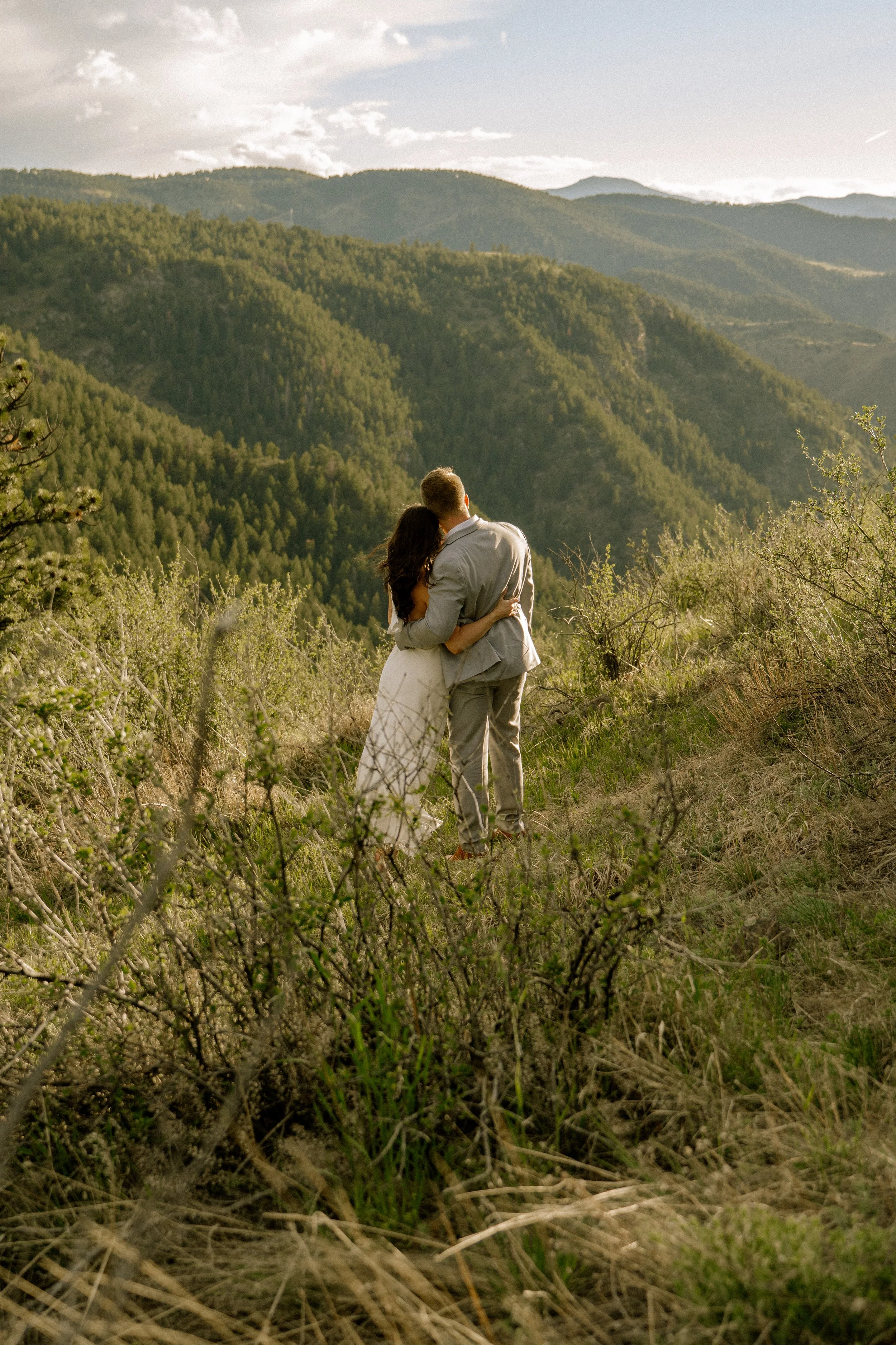 A couple standing close together, embracing and facing away from the camera, in a scenic mountain landscape with green hills and a cloudy sky.