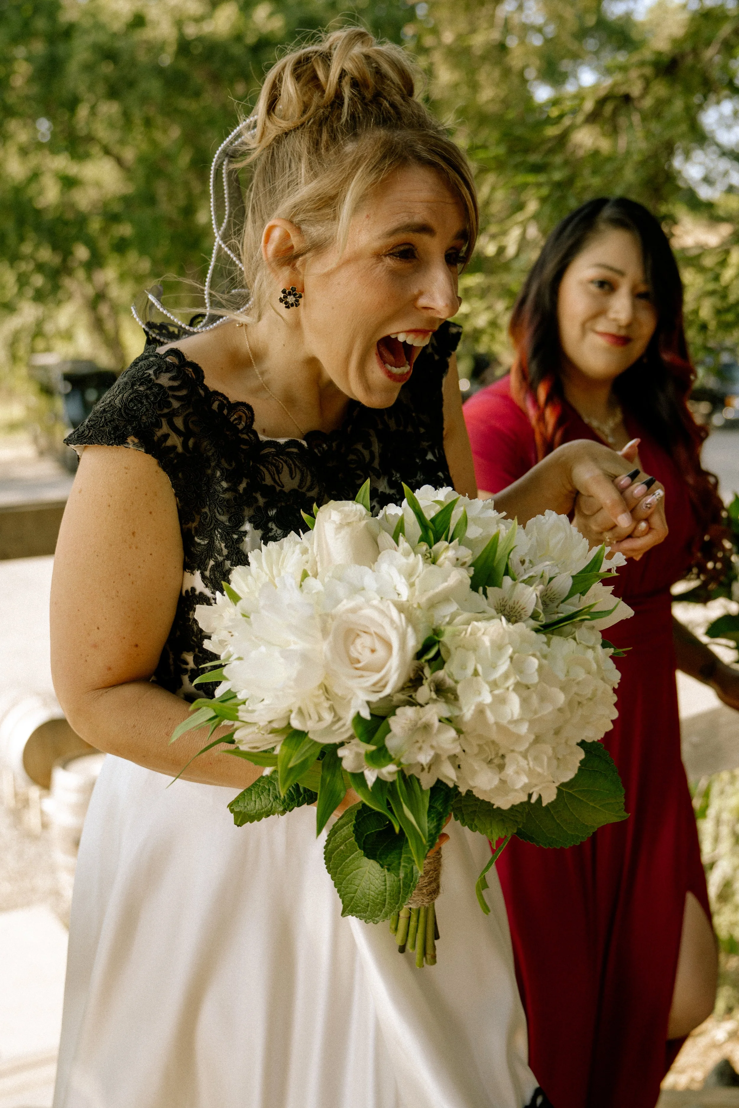 A woman in a black lace top and white skirt holding a large bouquet of white flowers while celebrating, with another woman in a red dress smiling in the background. They are outdoors with green trees.