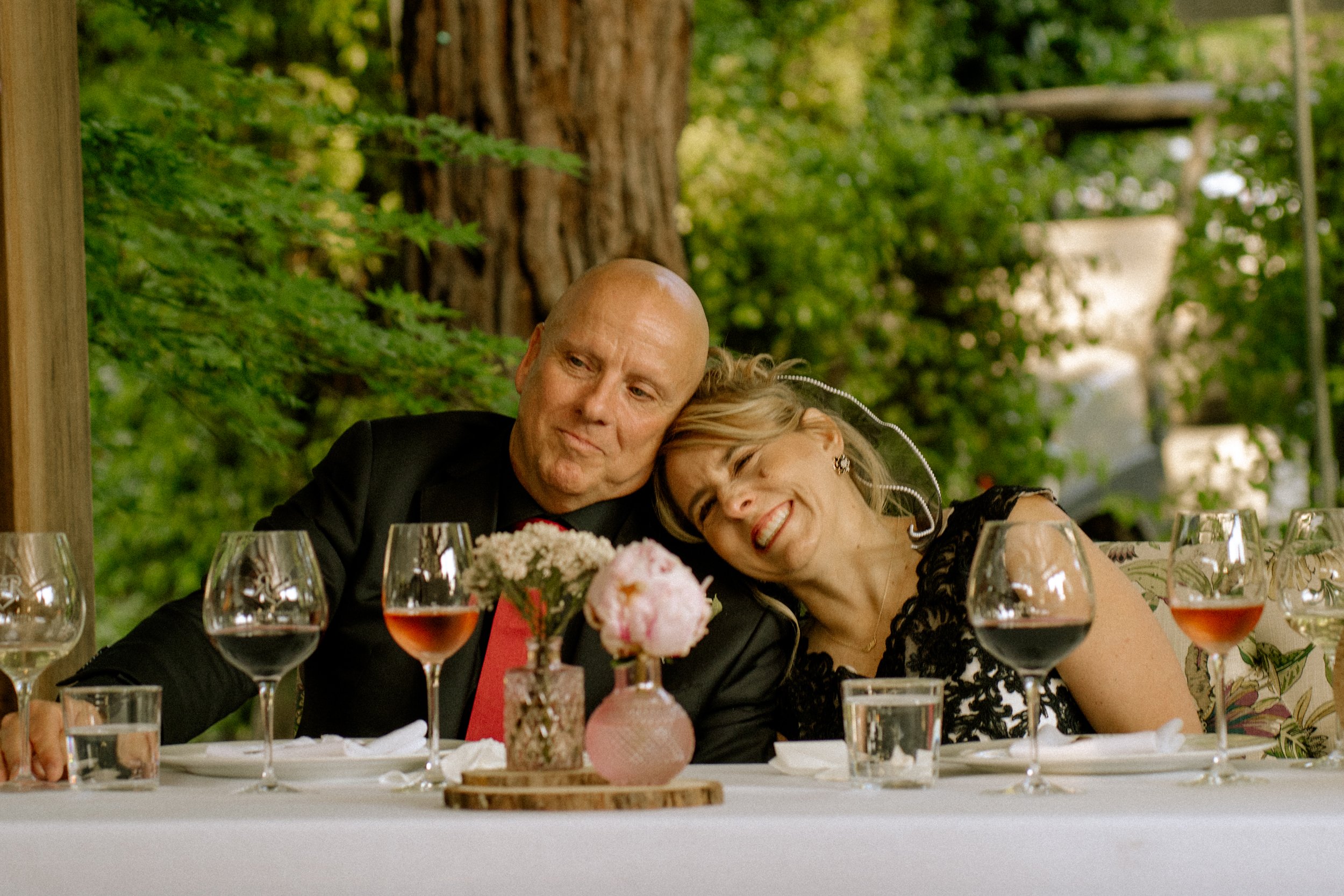 A man and a woman sitting together at a dining table, smiling and resting their heads on each other, surrounded by wine glasses and flowers, outdoors with green trees in the background.