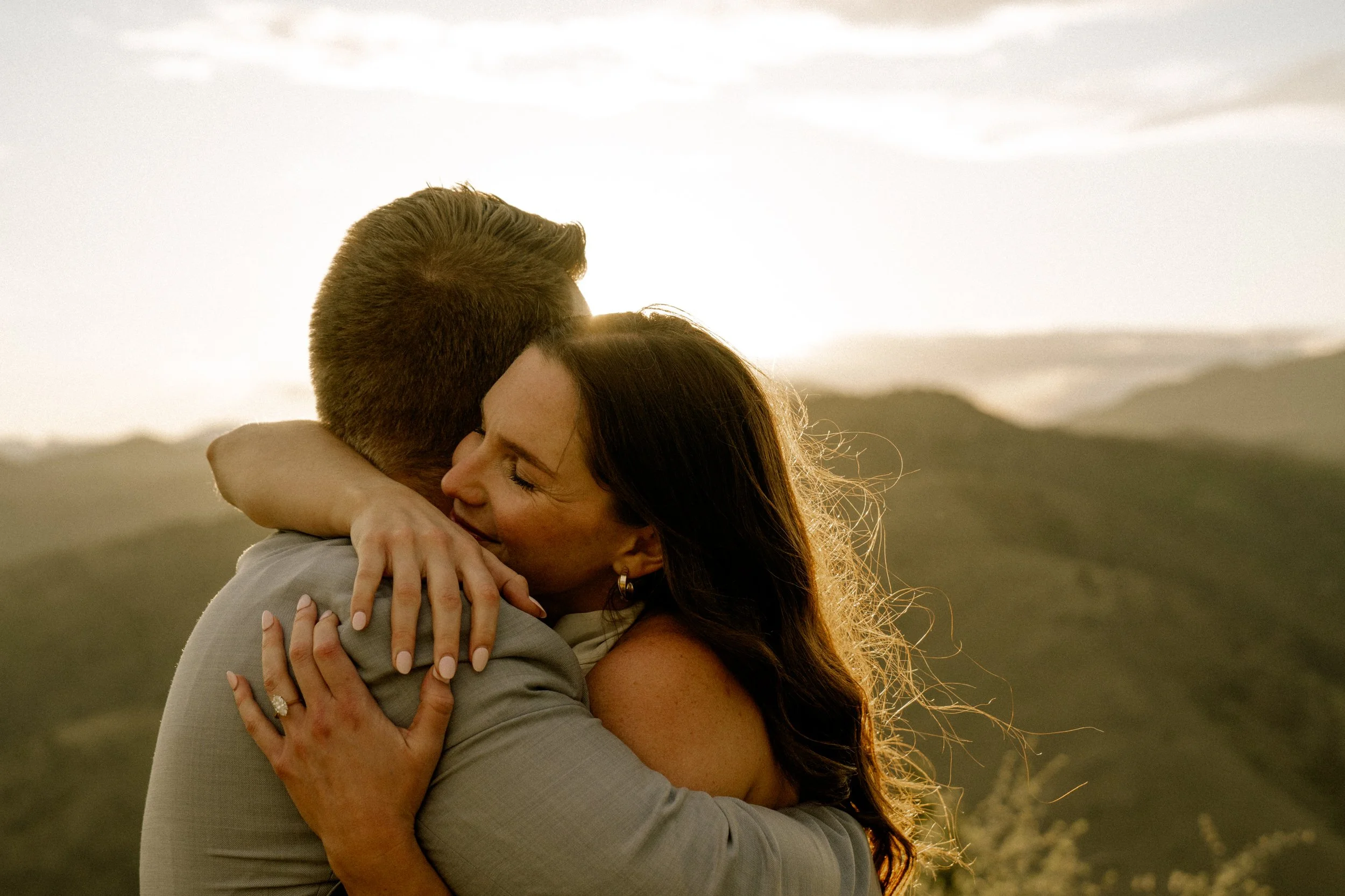 A man and a woman embrace outdoors at sunset, with hills and a cloudy sky in the background.