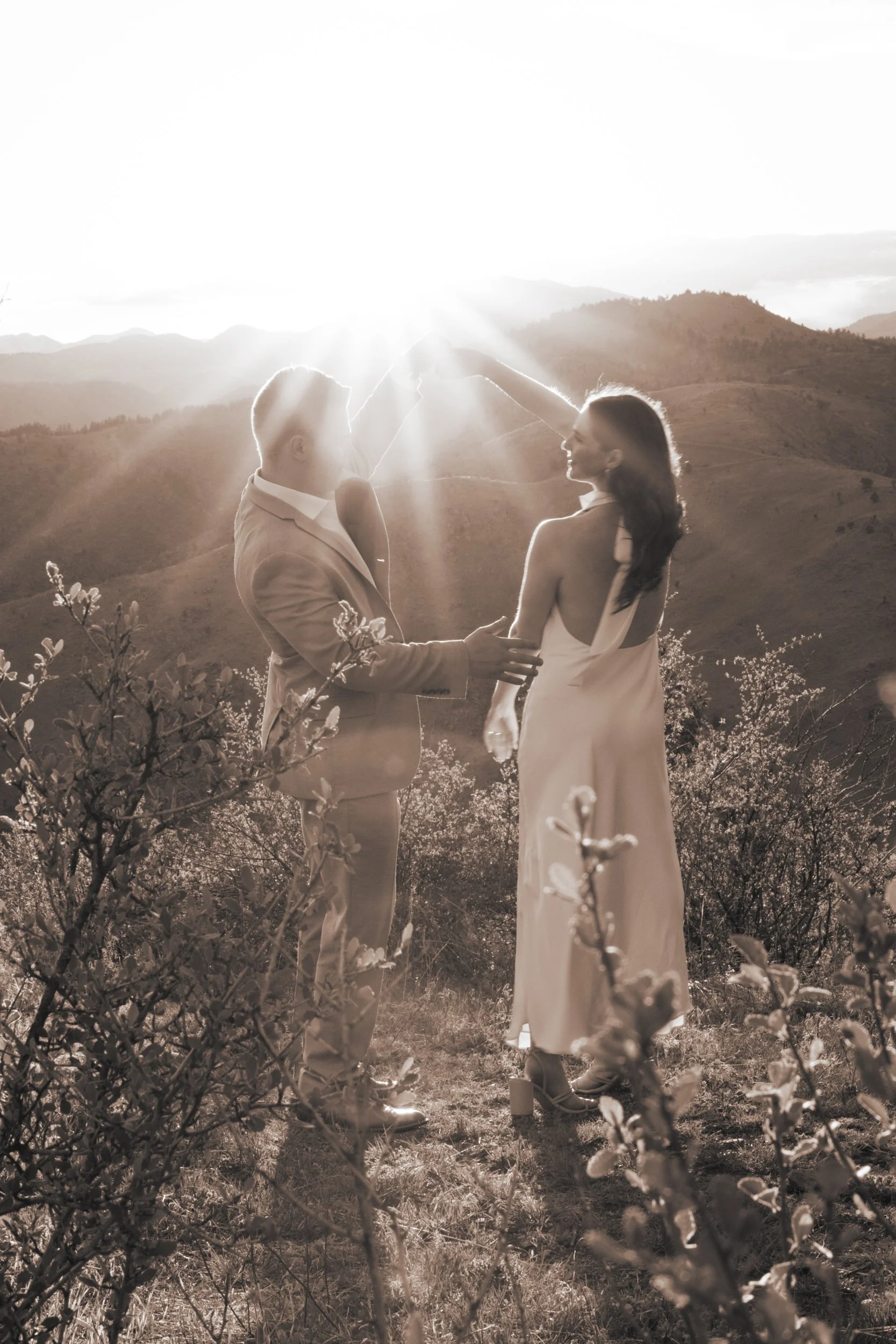 A couple in formal attire dancing outdoors on a hillside at sunset, with rolling hills visible in the background.