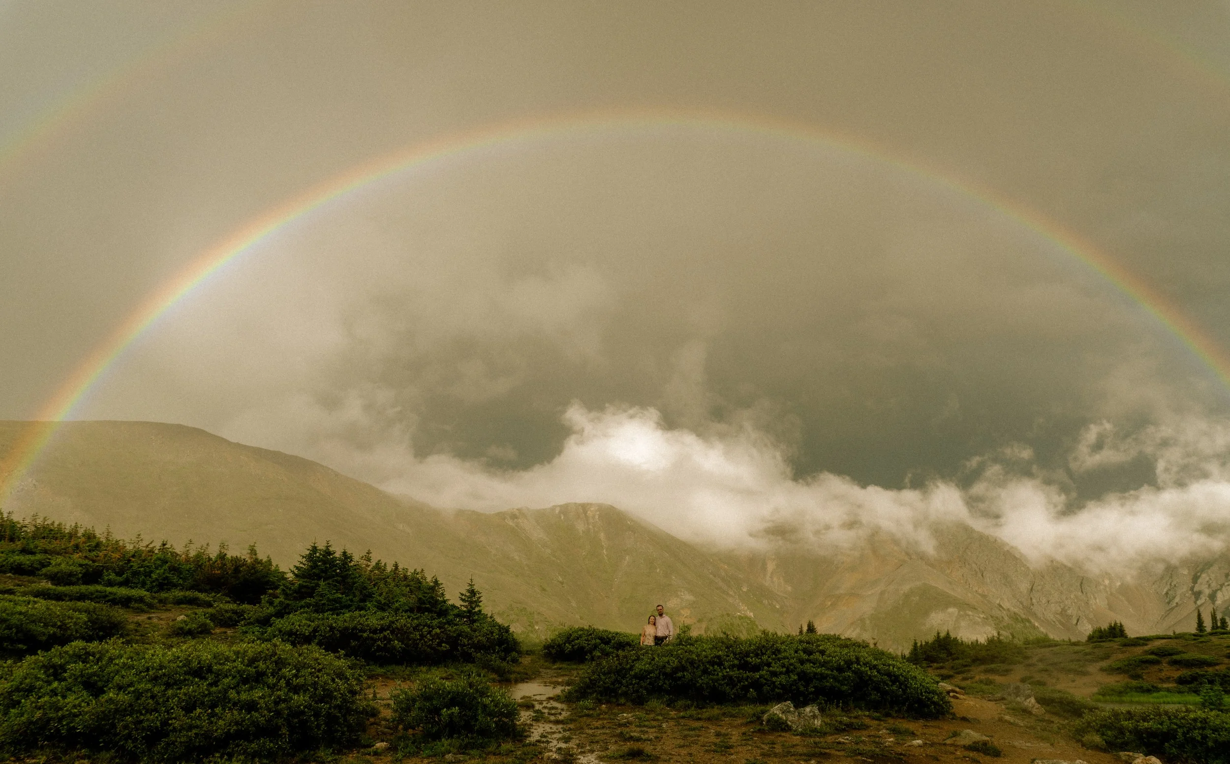 A landscape scene with a rainbow in the sky over green hills and trees, with two people standing in the foreground.