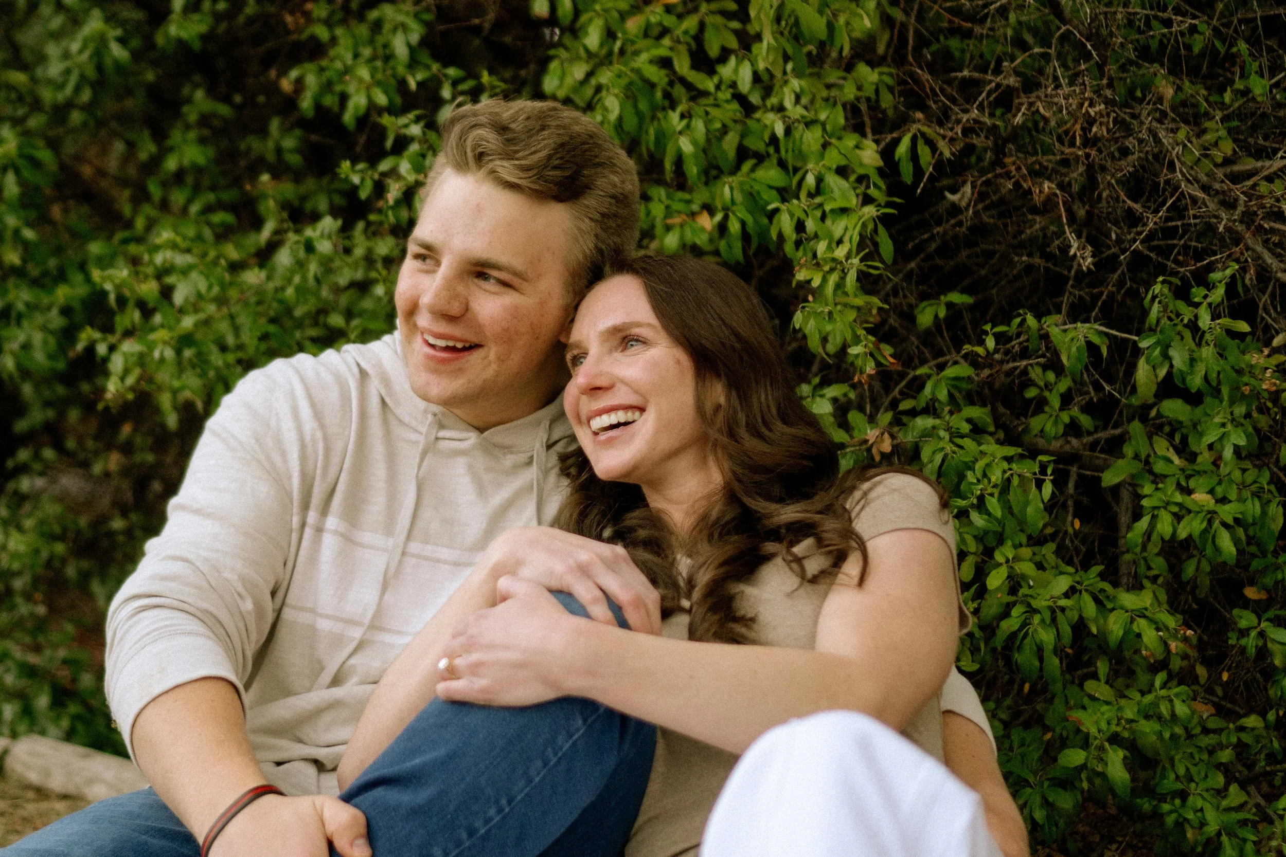 A young man and woman sitting closely together outdoors, smiling and leaning their heads against each other, surrounded by green foliage.
