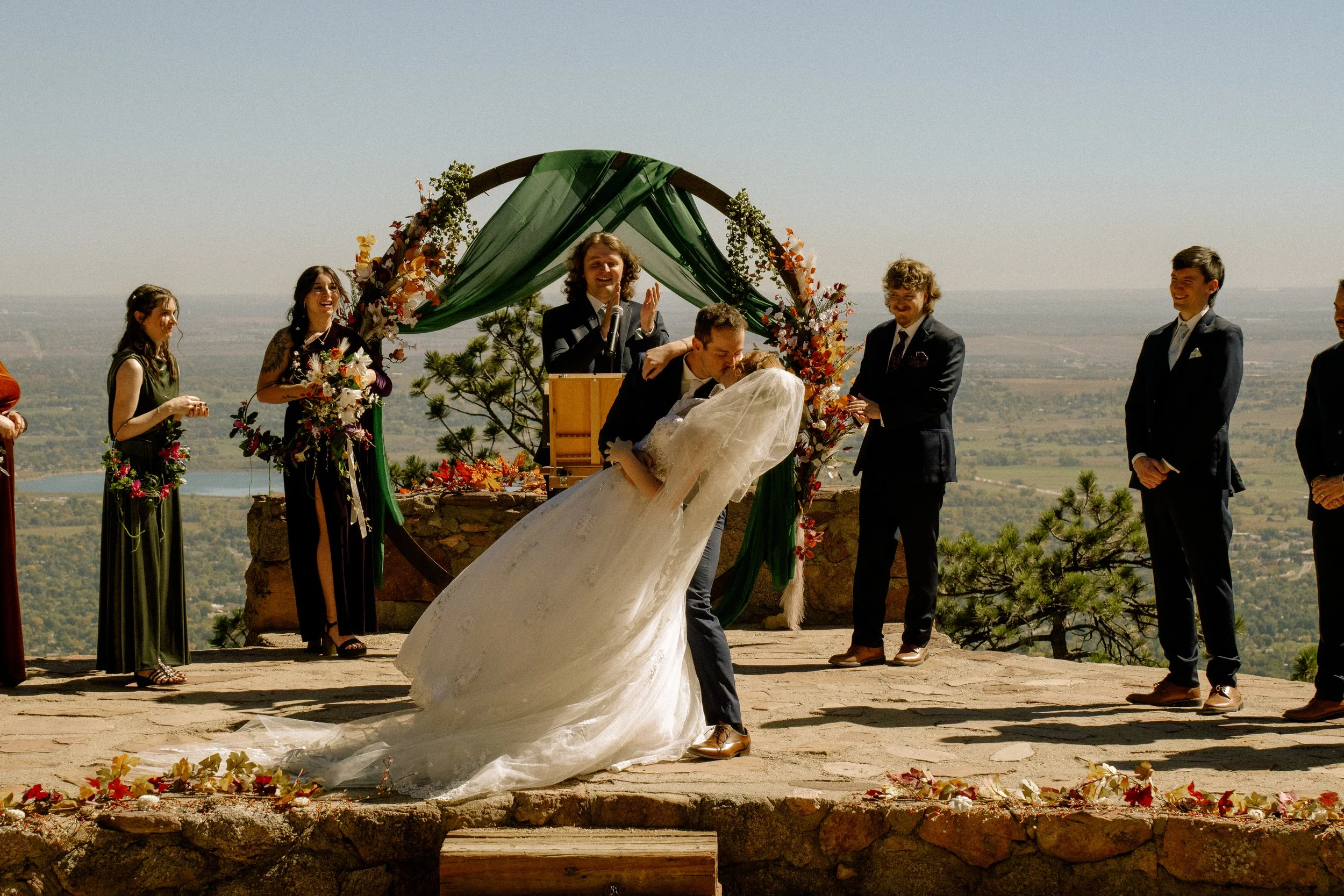 A couple gets married at an outdoor ceremony on a stone platform with scenic landscape in the background. The groom is kissing the bride, who is in a white wedding dress. Bridesmaids and groomsmen stand nearby, and an officiant stands behind a podium decorated with green fabric and flowers.