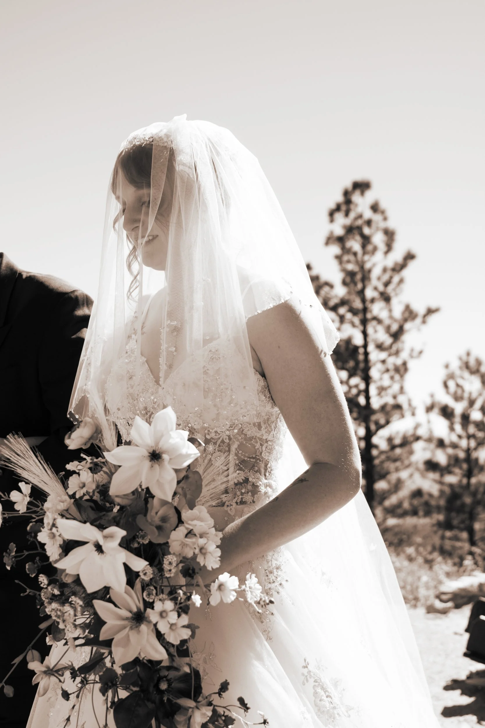 A bride in a wedding dress with a veil holding a bouquet of flowers, with trees and a clear sky in the background.