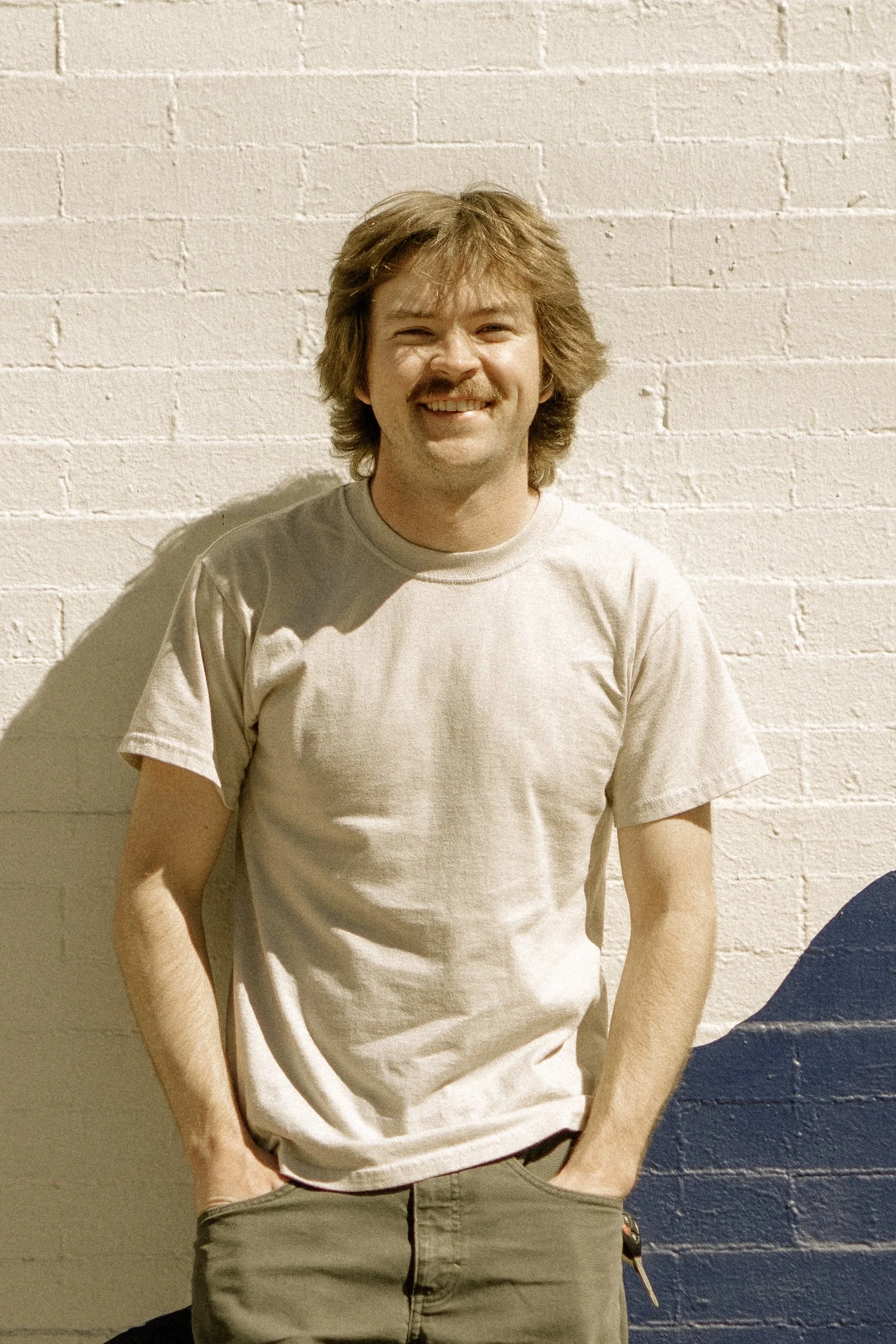 A man with brown curly hair, a mustache, and a goatee smiling, standing against a white brick wall with a dark blue section at the bottom. He is wearing a beige T-shirt and khaki pants.
