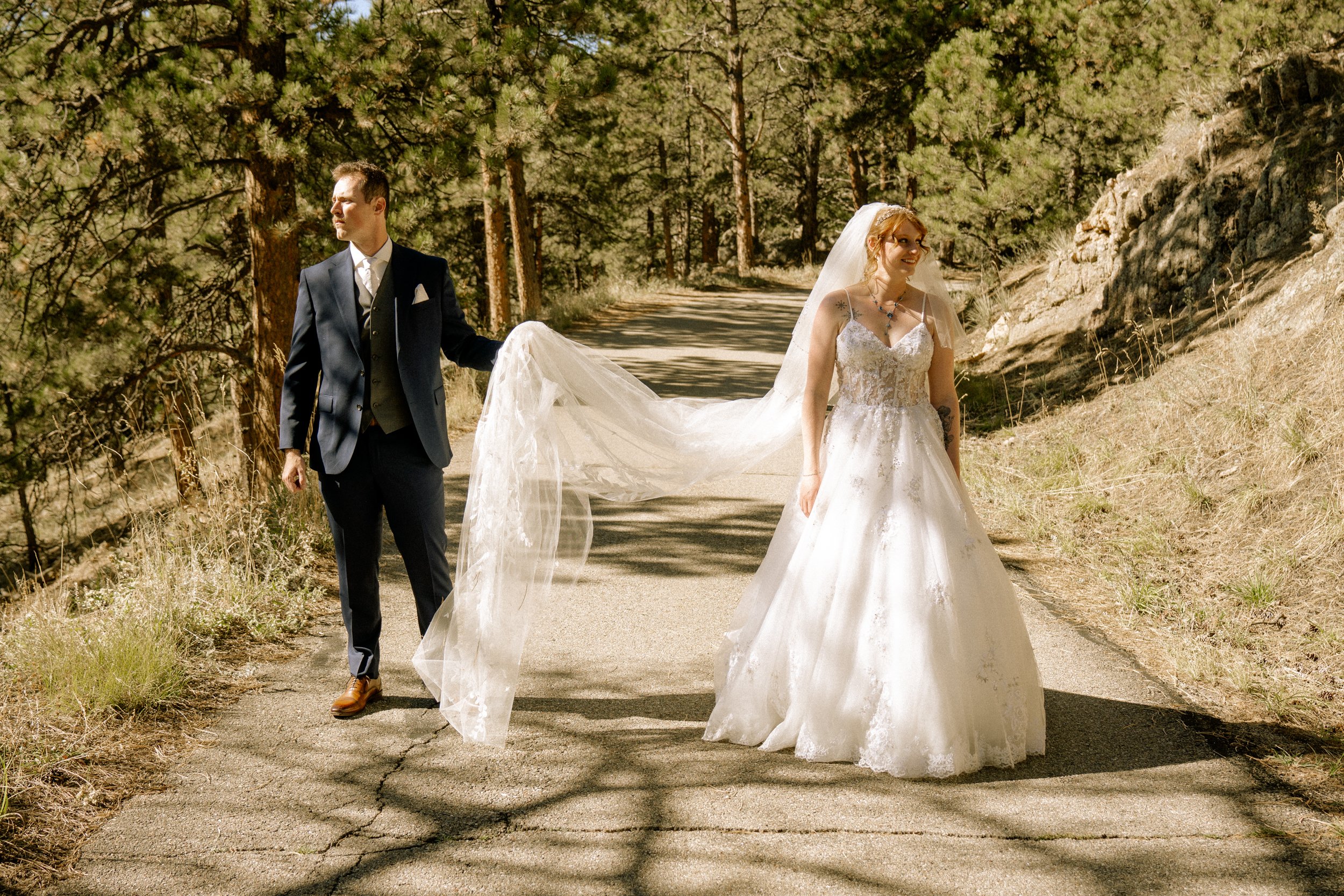 A bride and groom walking on a forest trail during a wedding photoshoot. The groom is holding the bride's train while she smiles. The scene is surrounded by trees and natural scenery.