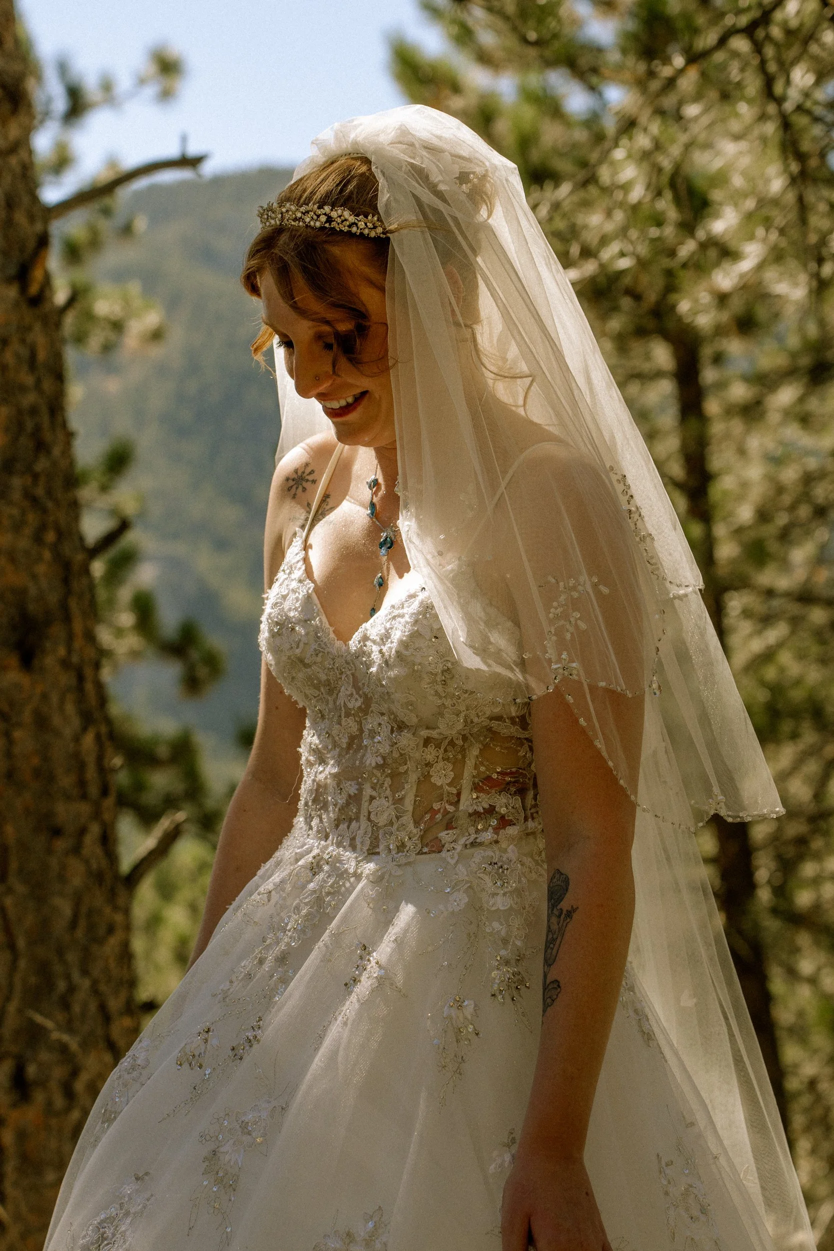 A smiling woman dressed in a wedding gown and veil, standing outdoors among trees with mountains in the background.