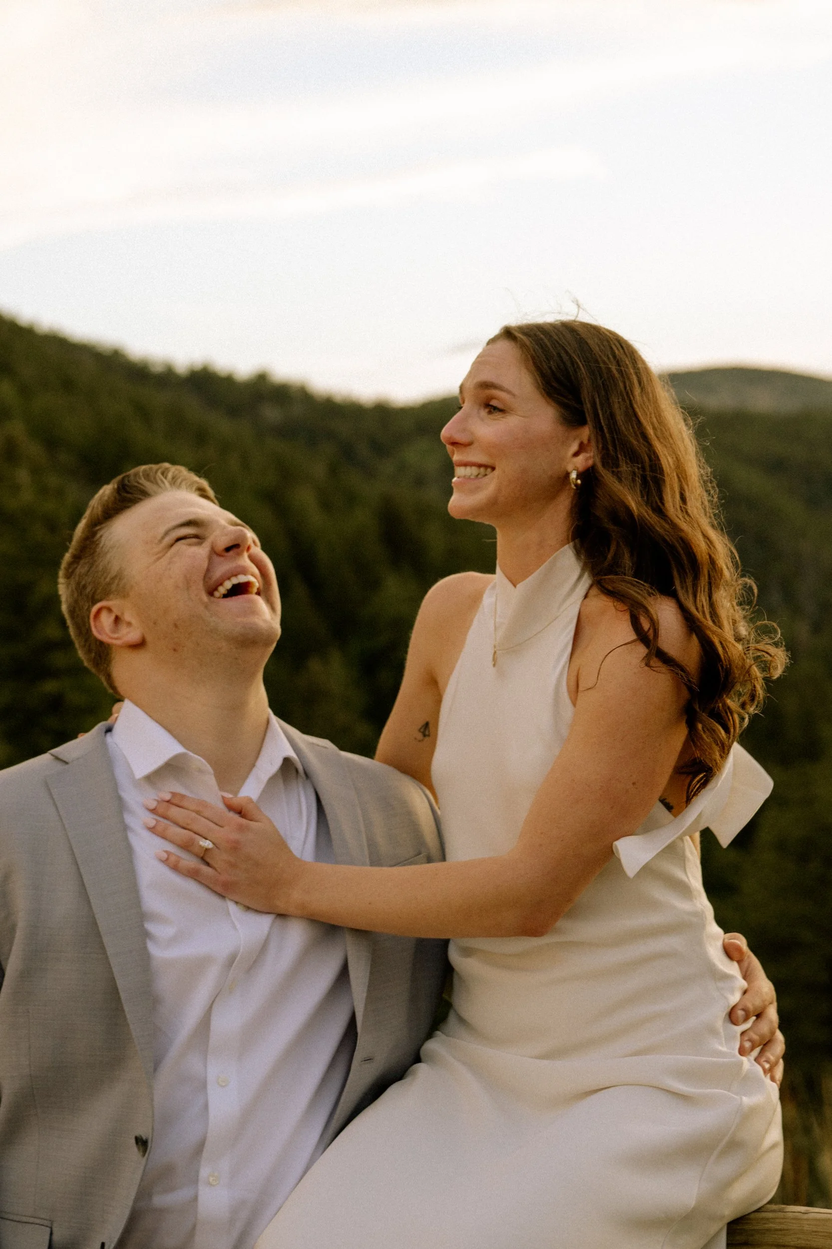 A happy couple dressed in wedding attire outdoors, smiling and laughing with a natural backdrop of mountains and trees during sunset.