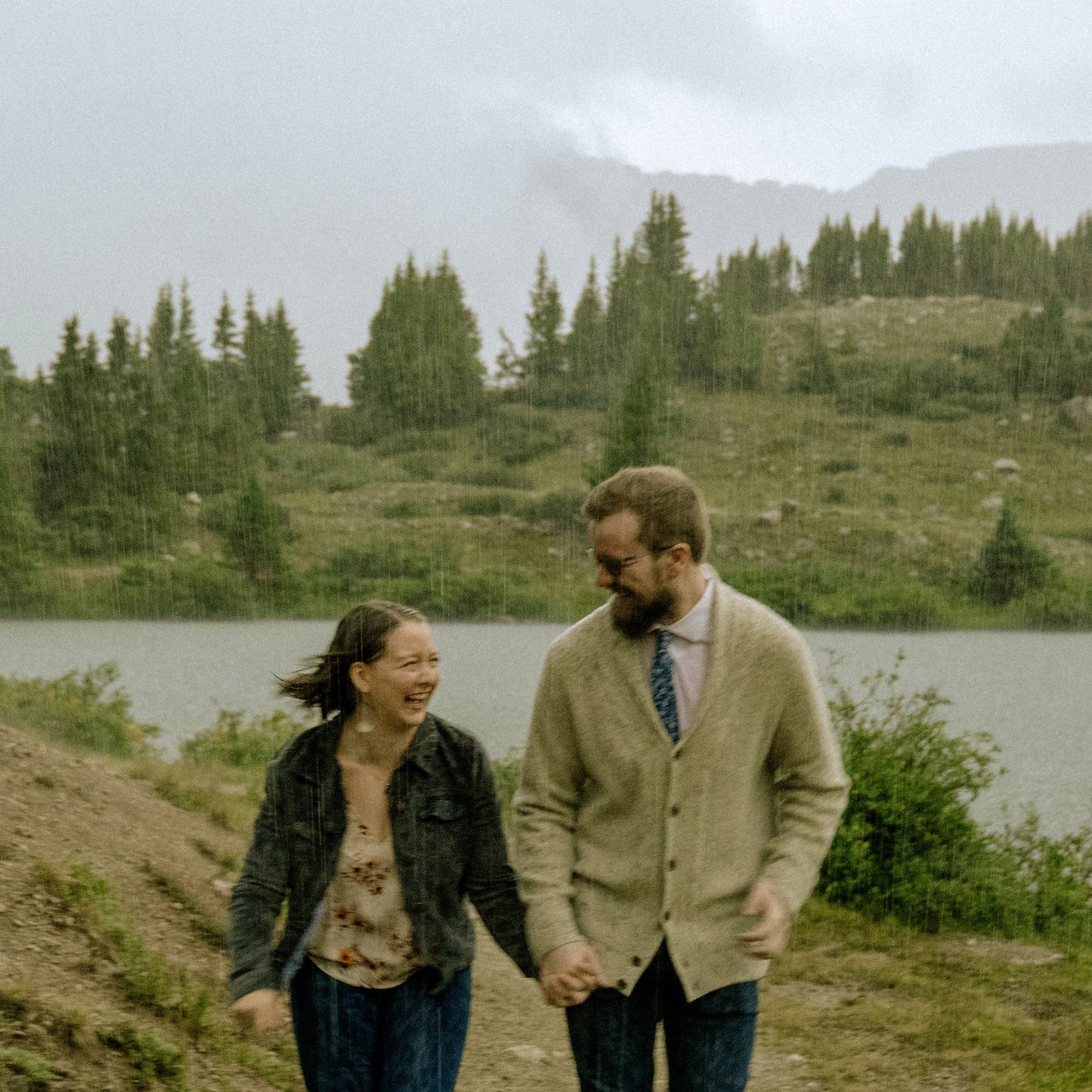 Two people holding hands walking outdoors in a natural setting with trees and mountainous hills in the background.
