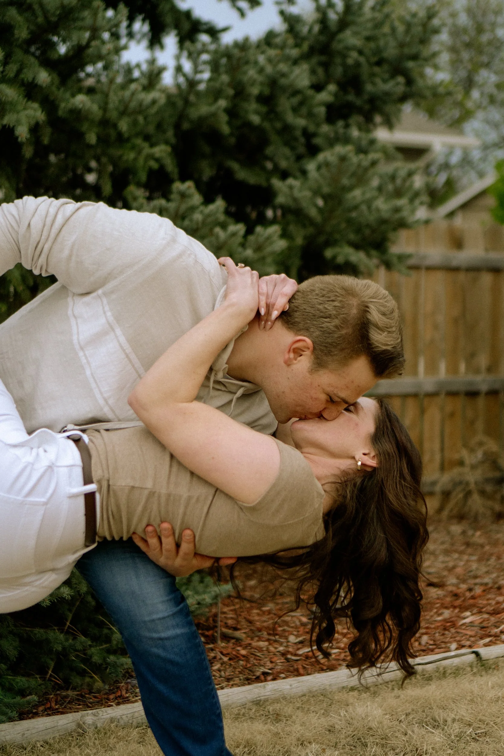 A couple sharing a kiss outdoors, with the woman bent backwards and the man leaning over her, in front of green trees and a wooden fence.