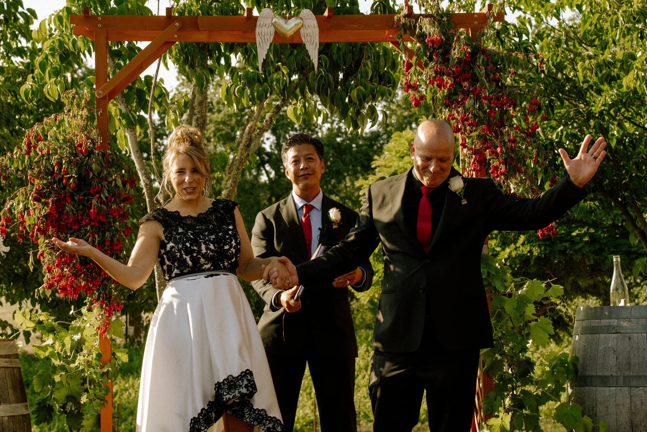 A wedding ceremony taking place outdoors under a wooden arch decorated with hanging red flowers and greenery. A bride in a white dress with black lace details and a groom in a black suit are holding hands, with an officiant standing behind them. The 