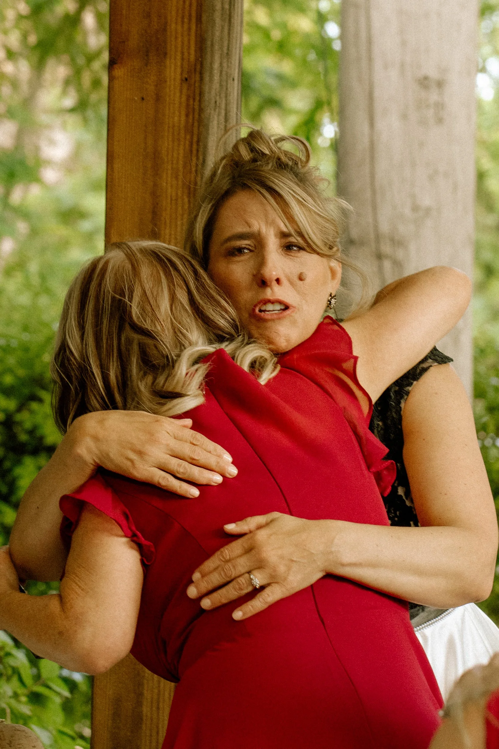 Two women embrace each other outdoors, with greenery and wooden posts in the background. One woman, with blonde hair and wearing a red dress, looks emotional.