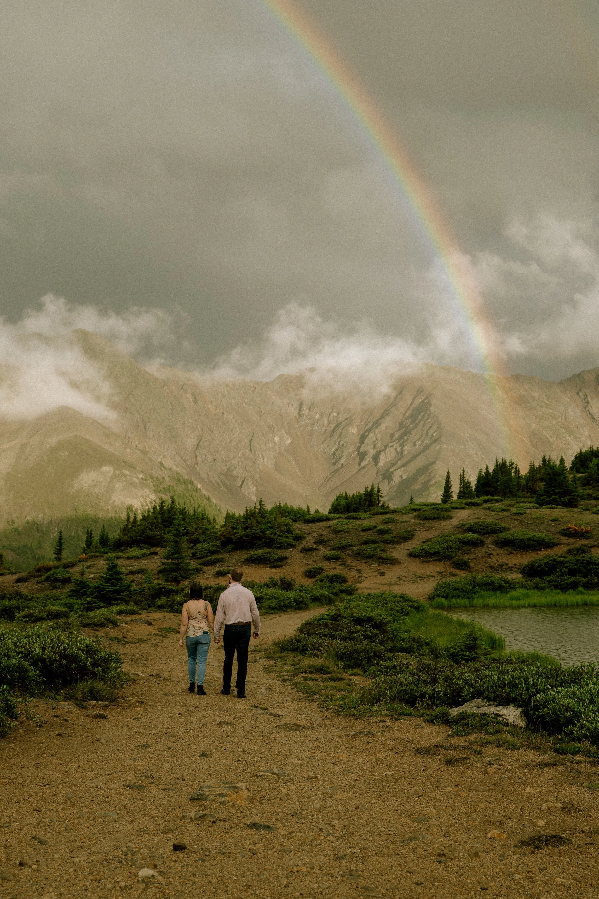 A couple walking hand in hand along a dirt path in a lush green landscape, with mountains and a rainbow in the cloudy sky overhead.