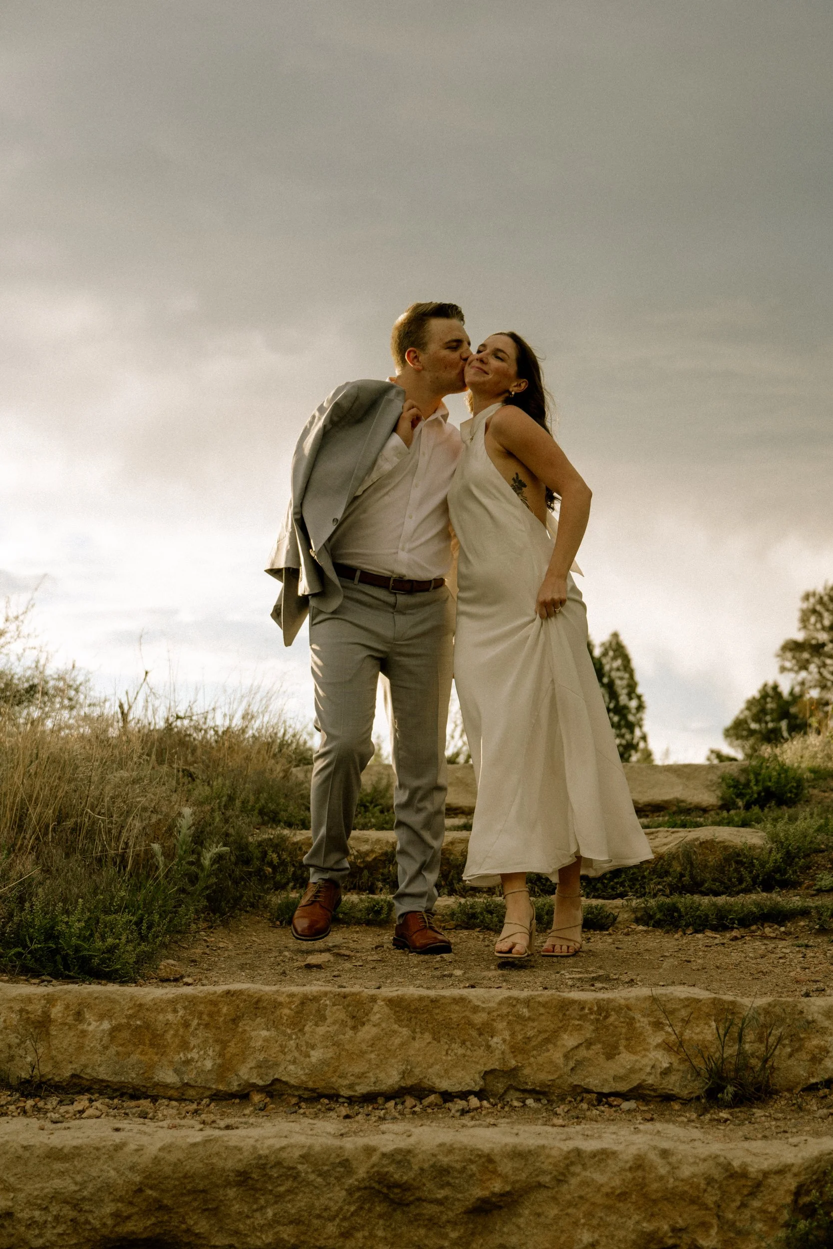 A couple standing outdoors on a rocky area, with the man kissing the woman on her cheek. The woman is wearing a long white dress and the man is in a white shirt, gray suit jacket, and gray pants, with a jacket draped over his shoulder. They are smili