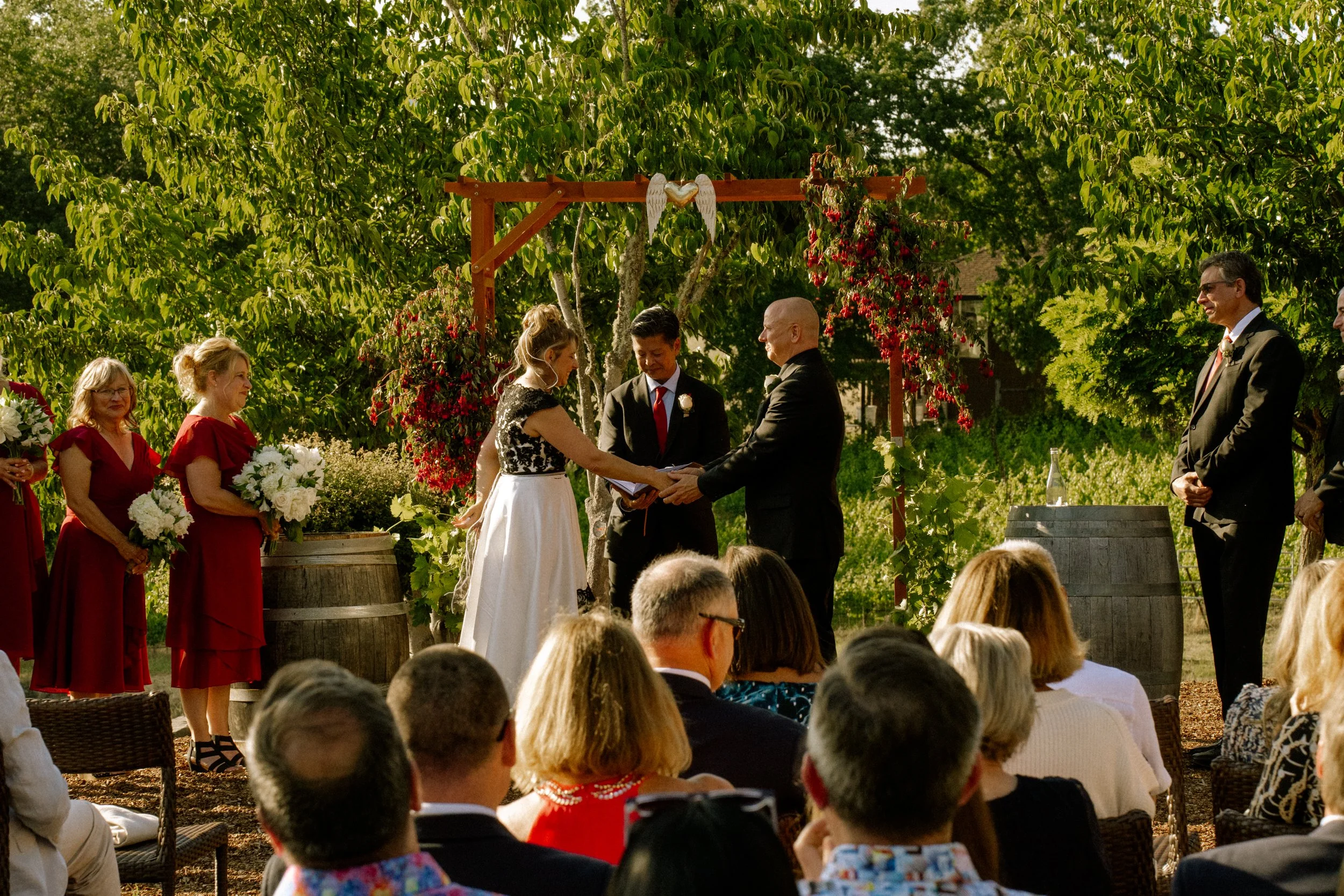 A group of people dressed in formal attire participate in an outdoor wedding ceremony. The bride and groom are exchanging vows under a wooden arch decorated with red flowers and a heart with angel wings. Guests are seated, and the background features