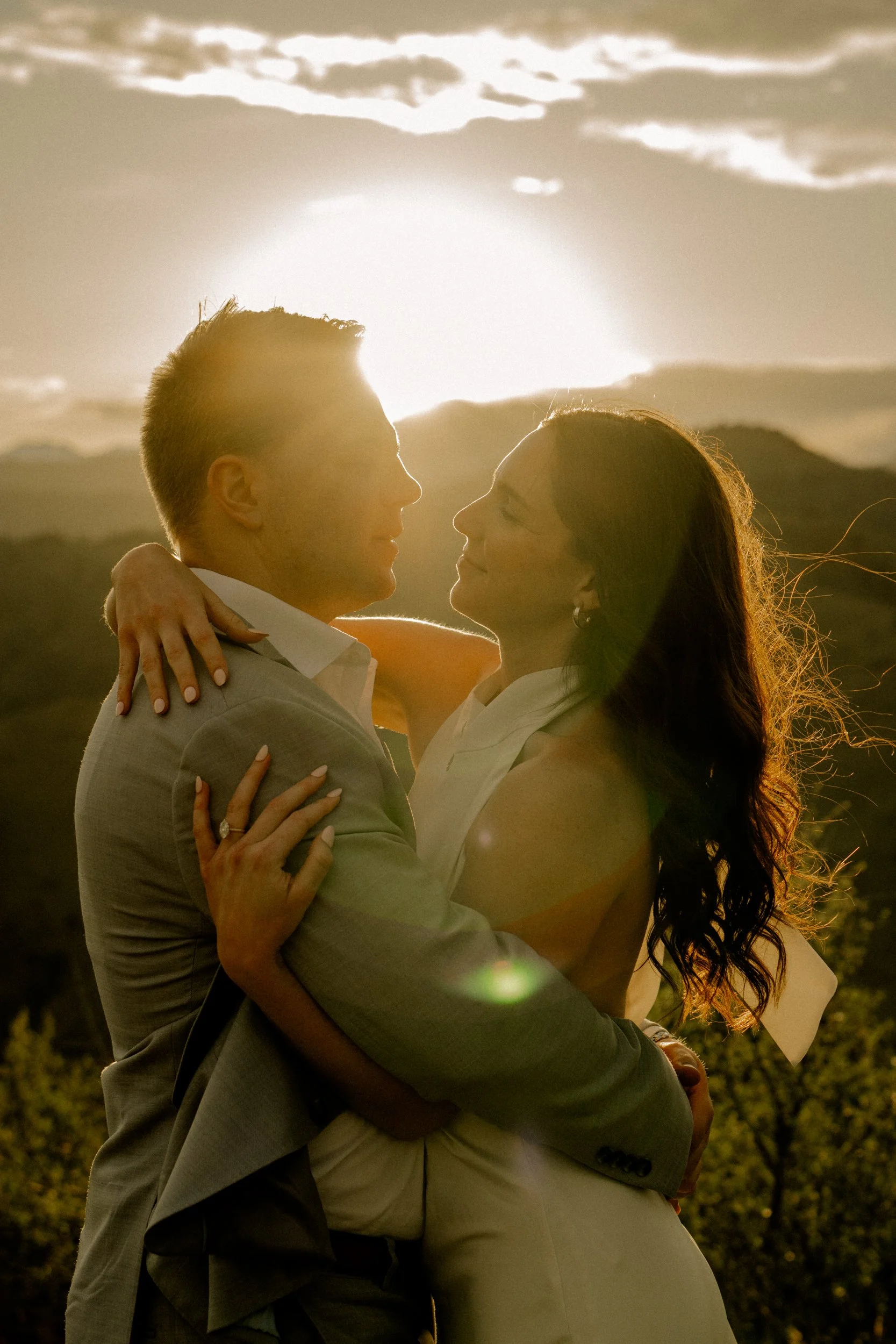 A couple embracing outdoors during sunset, with the sun setting behind them and creating a warm glow, hills or mountains in the background.