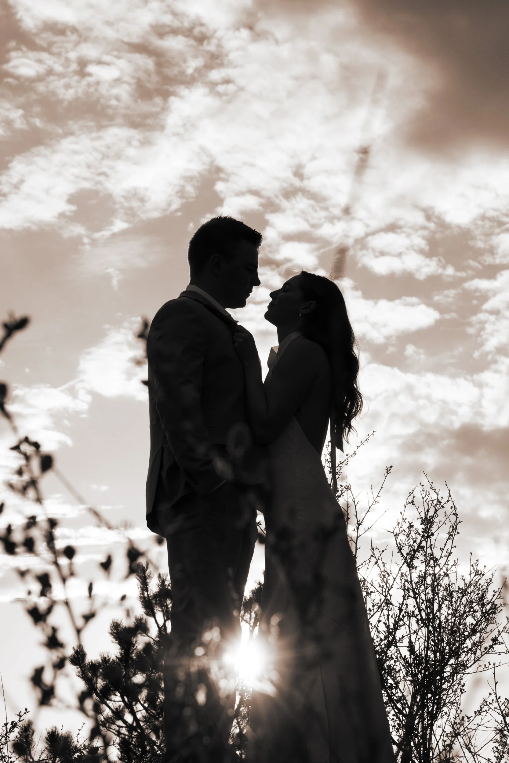 Silhouette of a man and woman facing each other outdoors against a cloudy sky, with the sun low behind trees in the foreground.