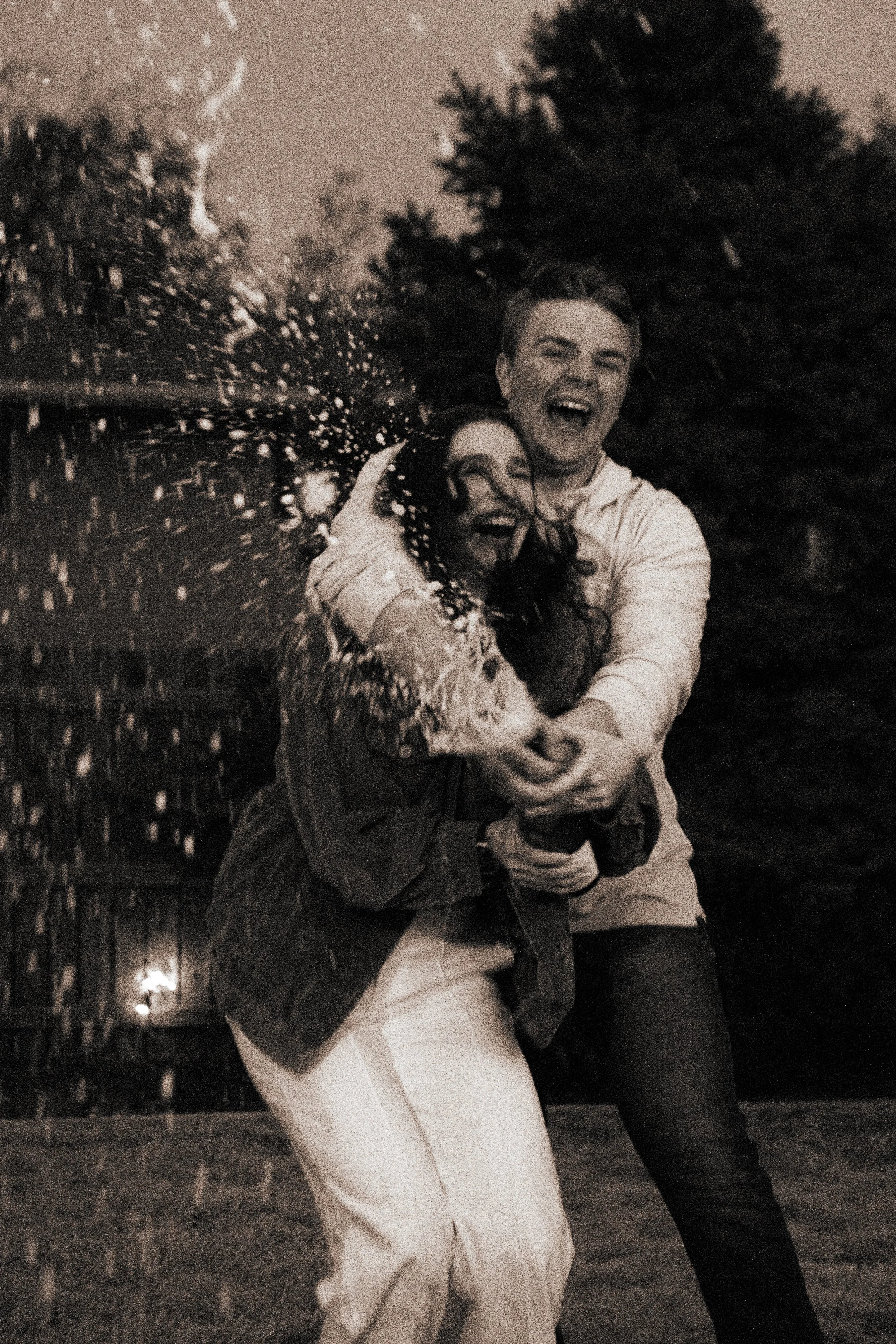 Two people, a man and a woman, are joyfully celebrating outdoors at night with a garden or park in the background. They are smiling and embracing each other.