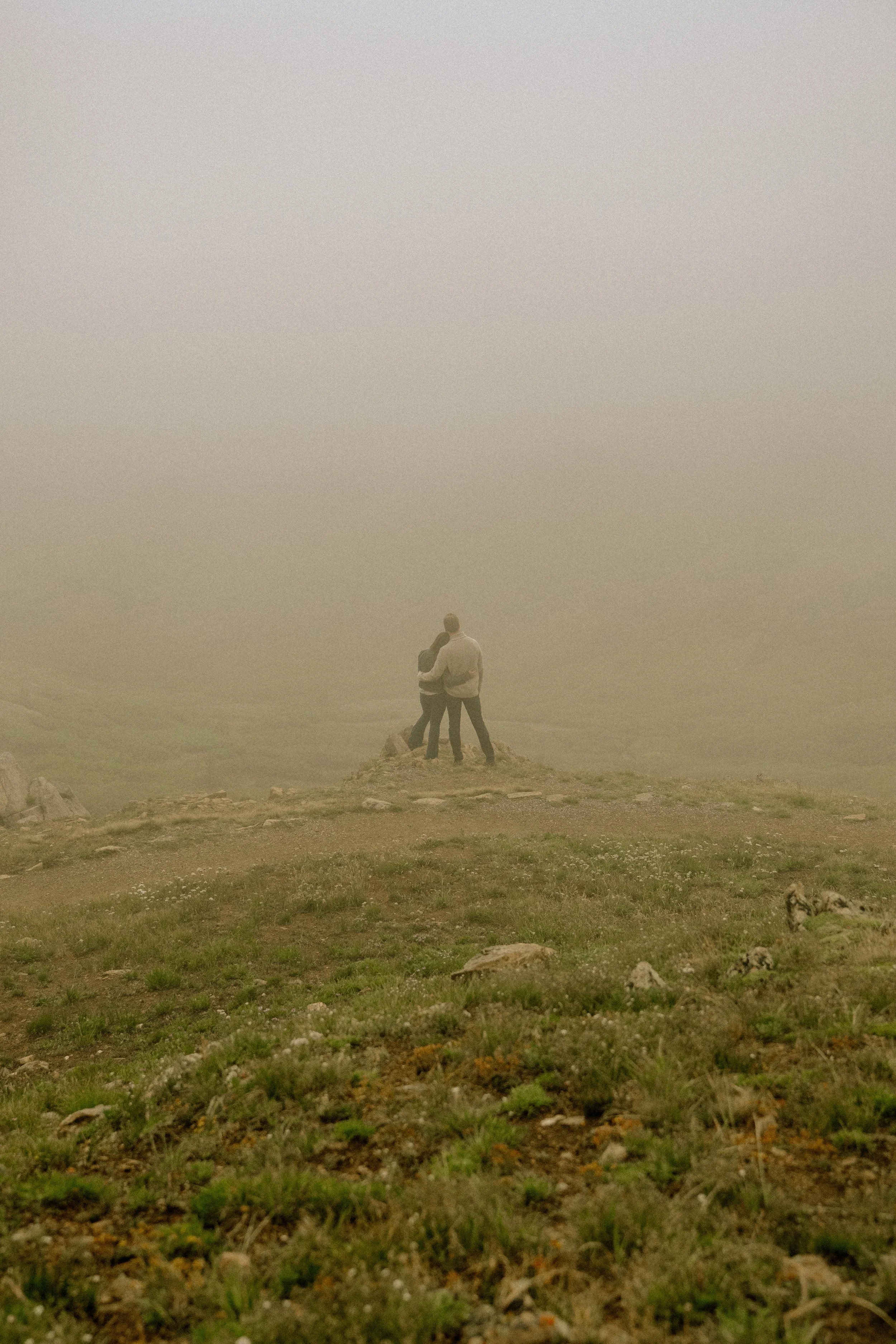 Two people standing on a grassy hillside with rocks, foggy weather obscuring the background, and a dog lying in the grass.