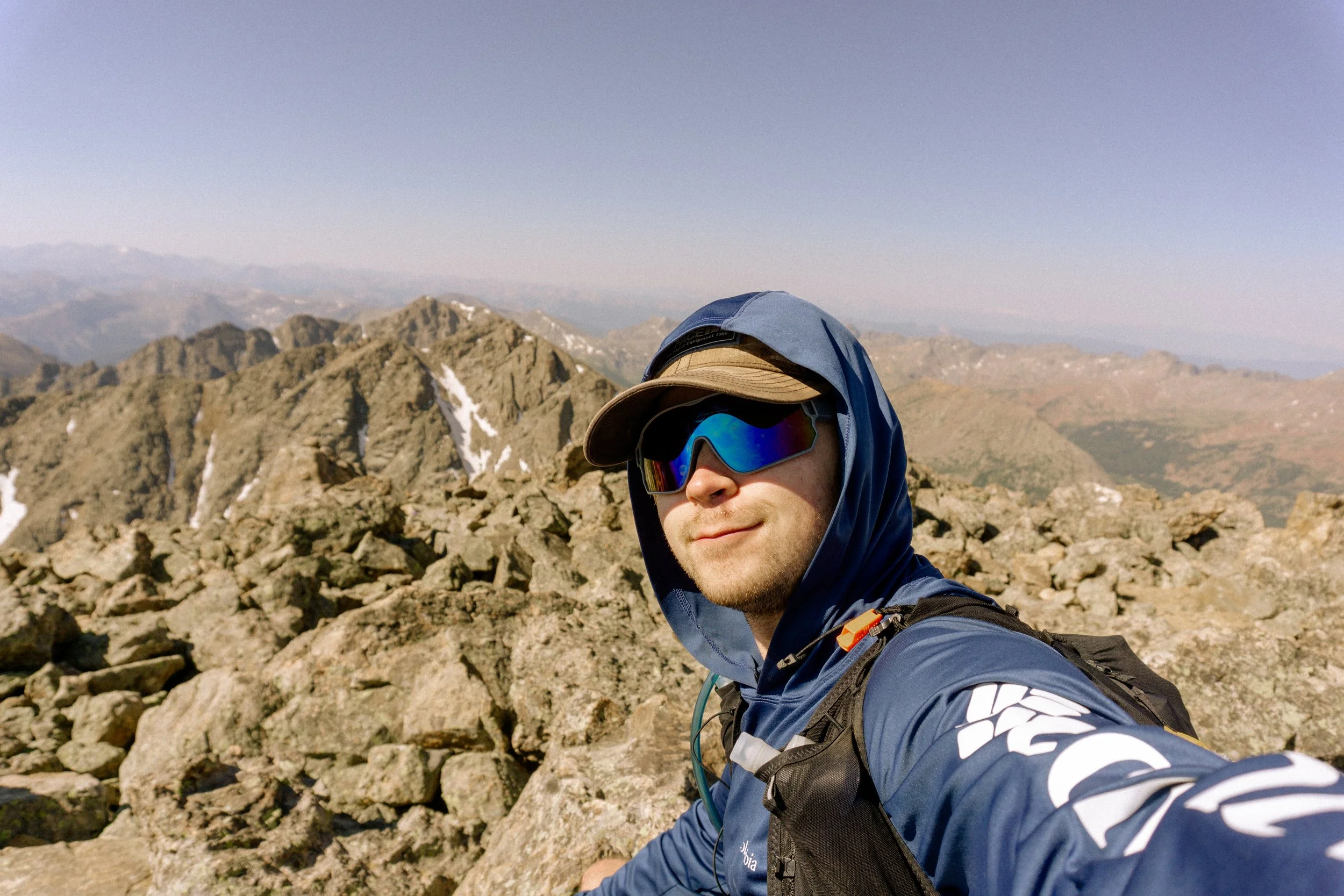 A man taking a selfie on a rocky mountain summit with mountains in the background, wearing sunglasses, a hat, and outdoor gear.