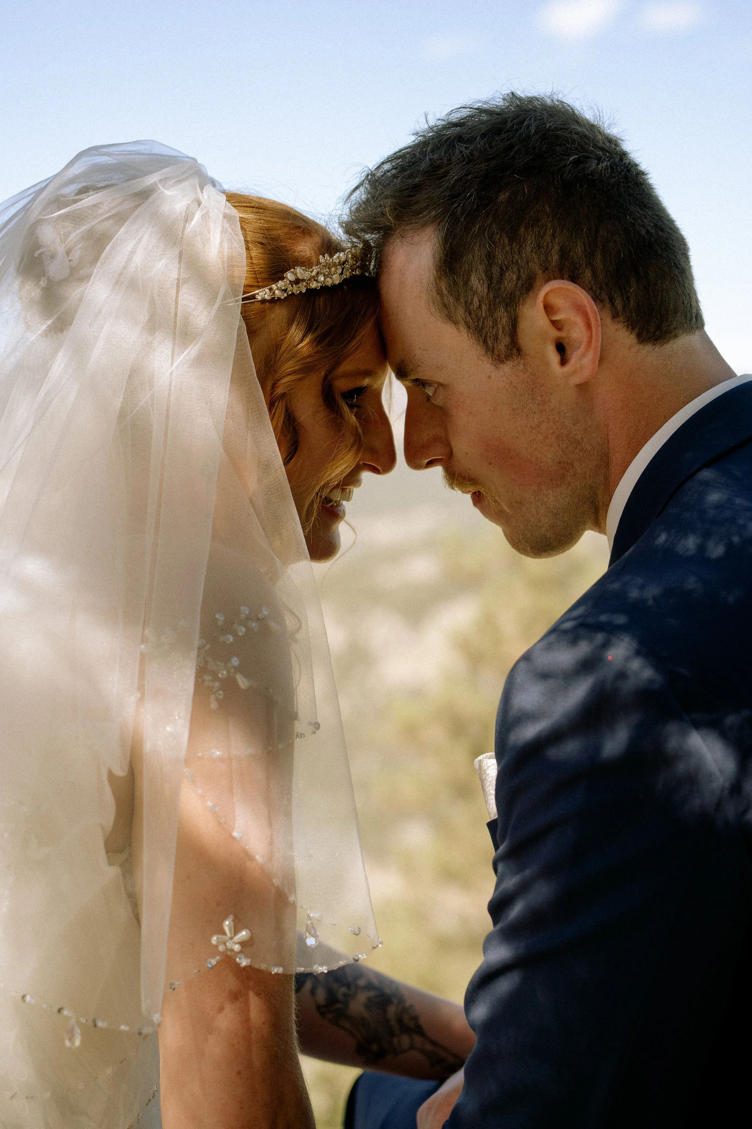 A bride and groom with foreheads touching, eyes closed, outdoors with a blurred background.